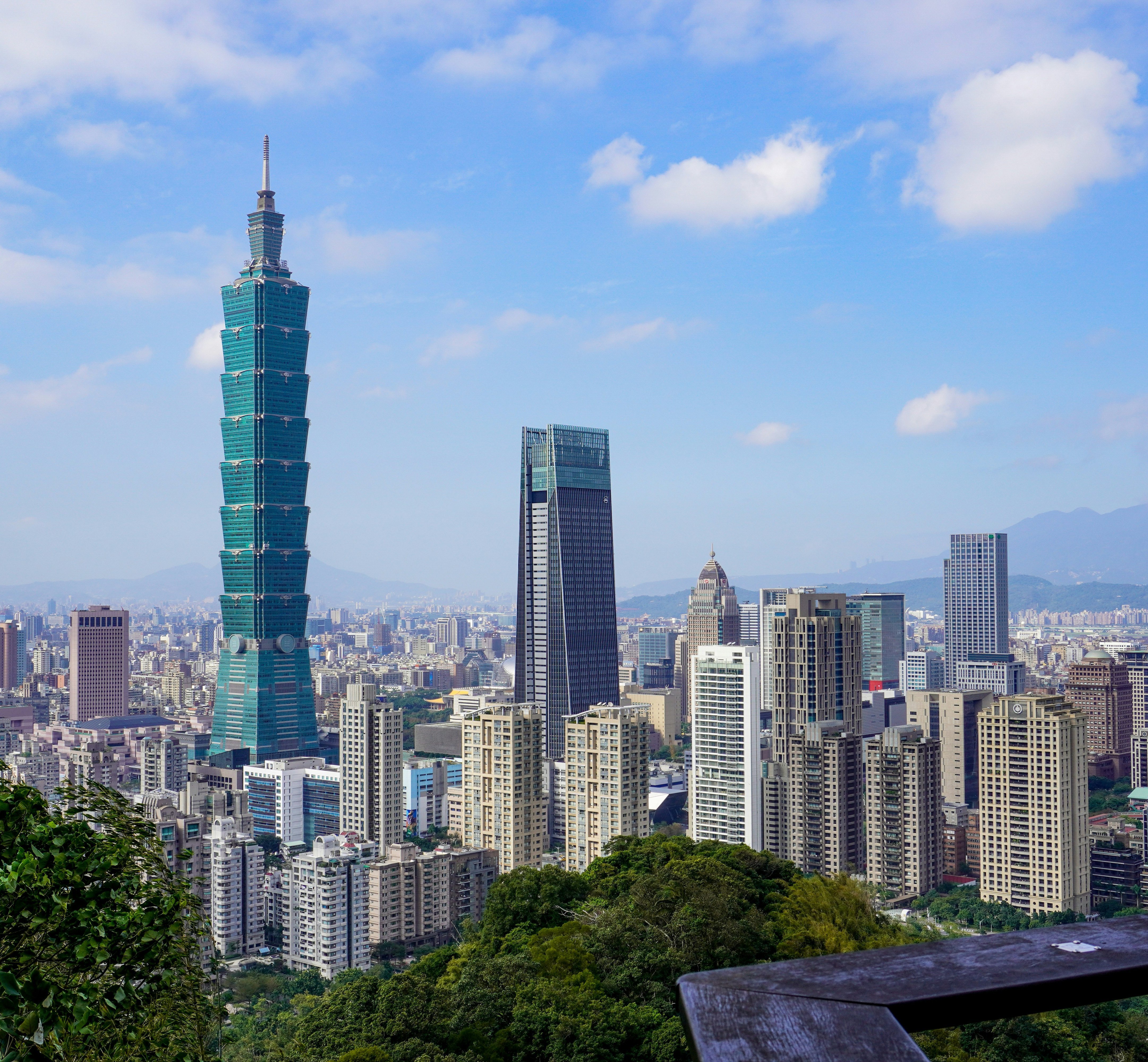 Die Skyline von Taipeh mit dem hohen Wolkenkratzer Taipeh 101, modernen Gebäuden und üppig grünen Bäumen im Vordergrund unter einem blauen Himmel mit vereinzelten Wolken.