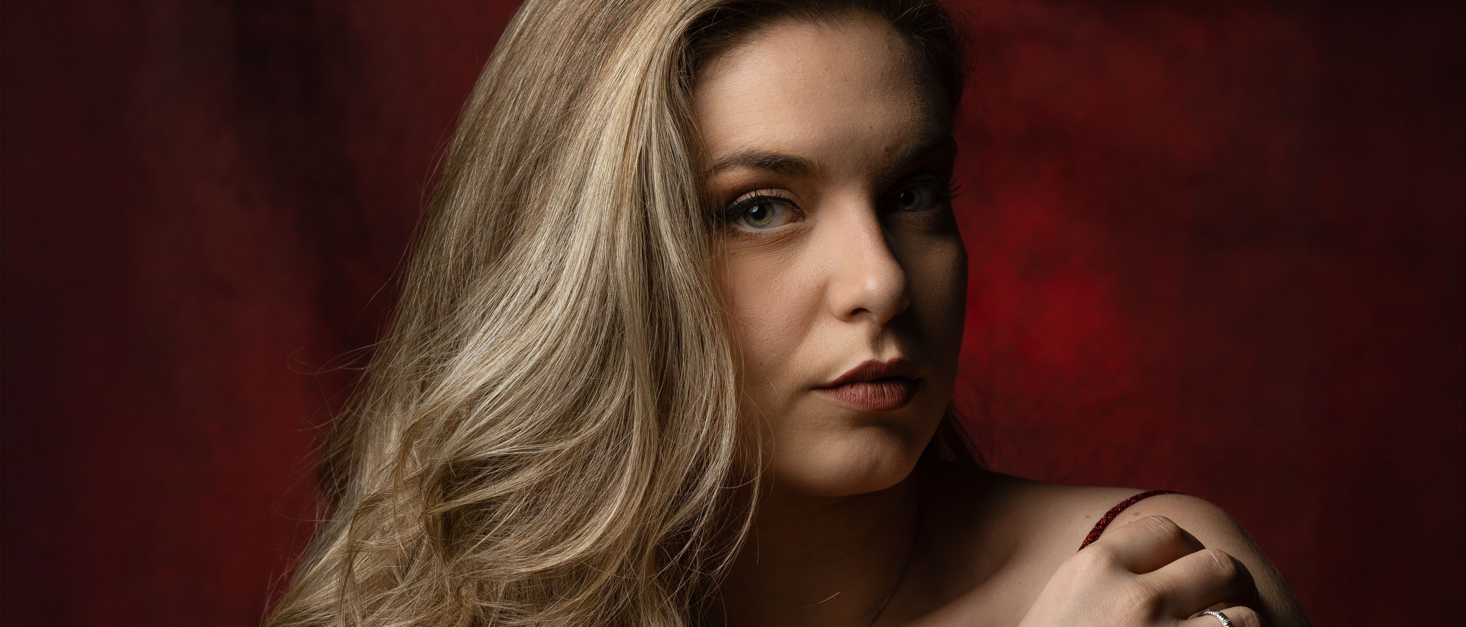 Eine Frau mit langem, gewelltem blondem Haar trägt ein ärmelloses, tiefrotes Kleid und Schmuck und posiert vor einem dunkelroten, strukturierten Hintergrund, während sie ihre Schulter berührt und in die Kamera schaut.