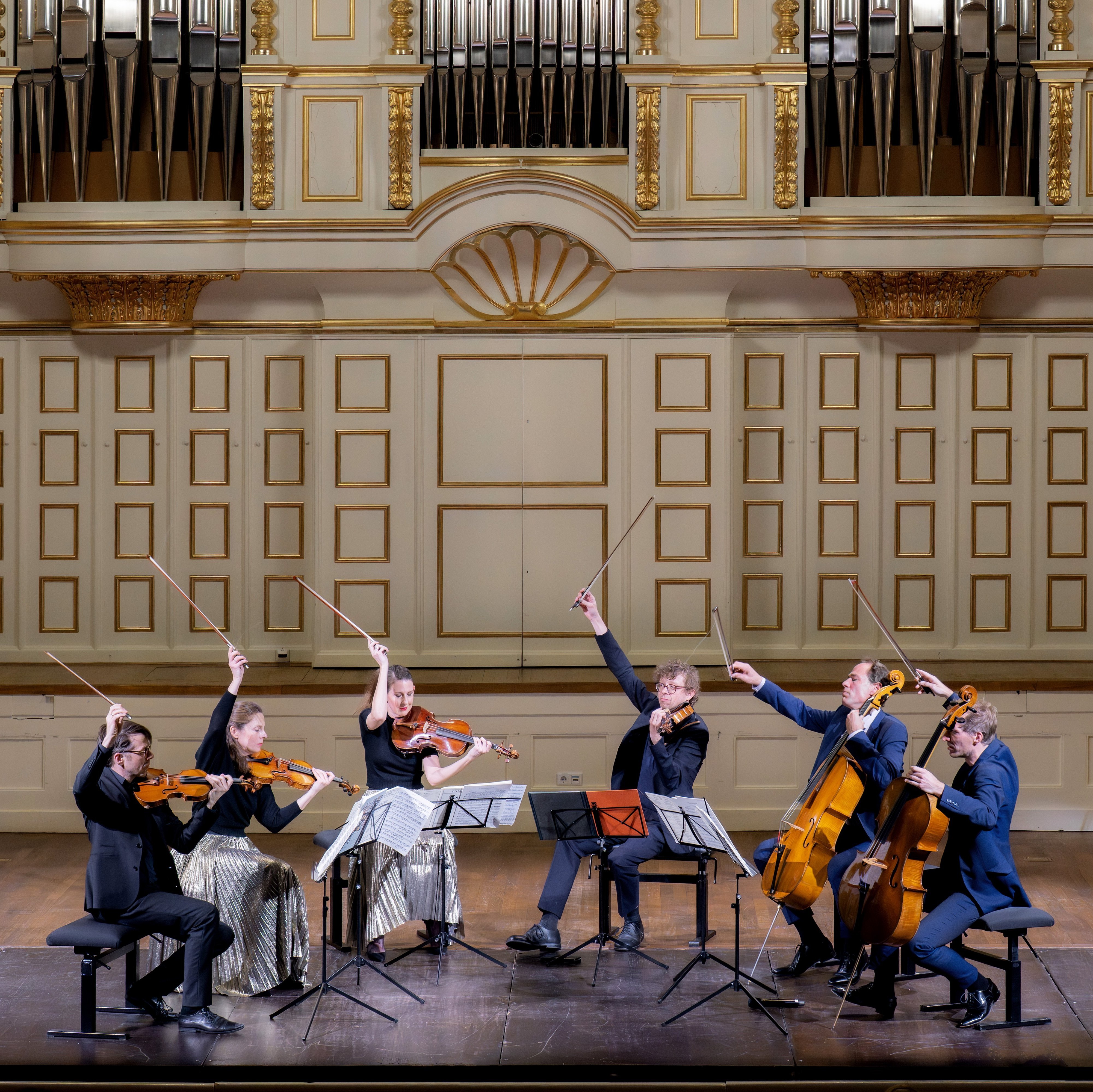Sechs Musiker mit Streichinstrumenten treten auf der Bühne eines prunkvollen Konzertsaals auf, heben ihre Bögen dramatisch in die Luft, vor ihnen stehen Notenständer, dahinter eine große Orgel.