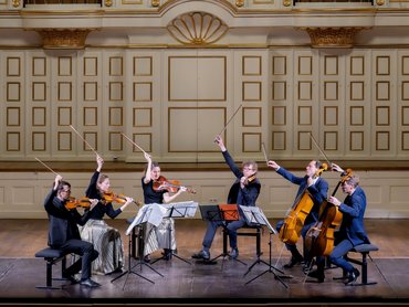 Sechs Musiker mit Streichinstrumenten treten auf der Bühne eines prunkvollen Konzertsaals auf, heben ihre Bögen dramatisch in die Luft, vor ihnen stehen Notenständer, dahinter eine große Orgel.