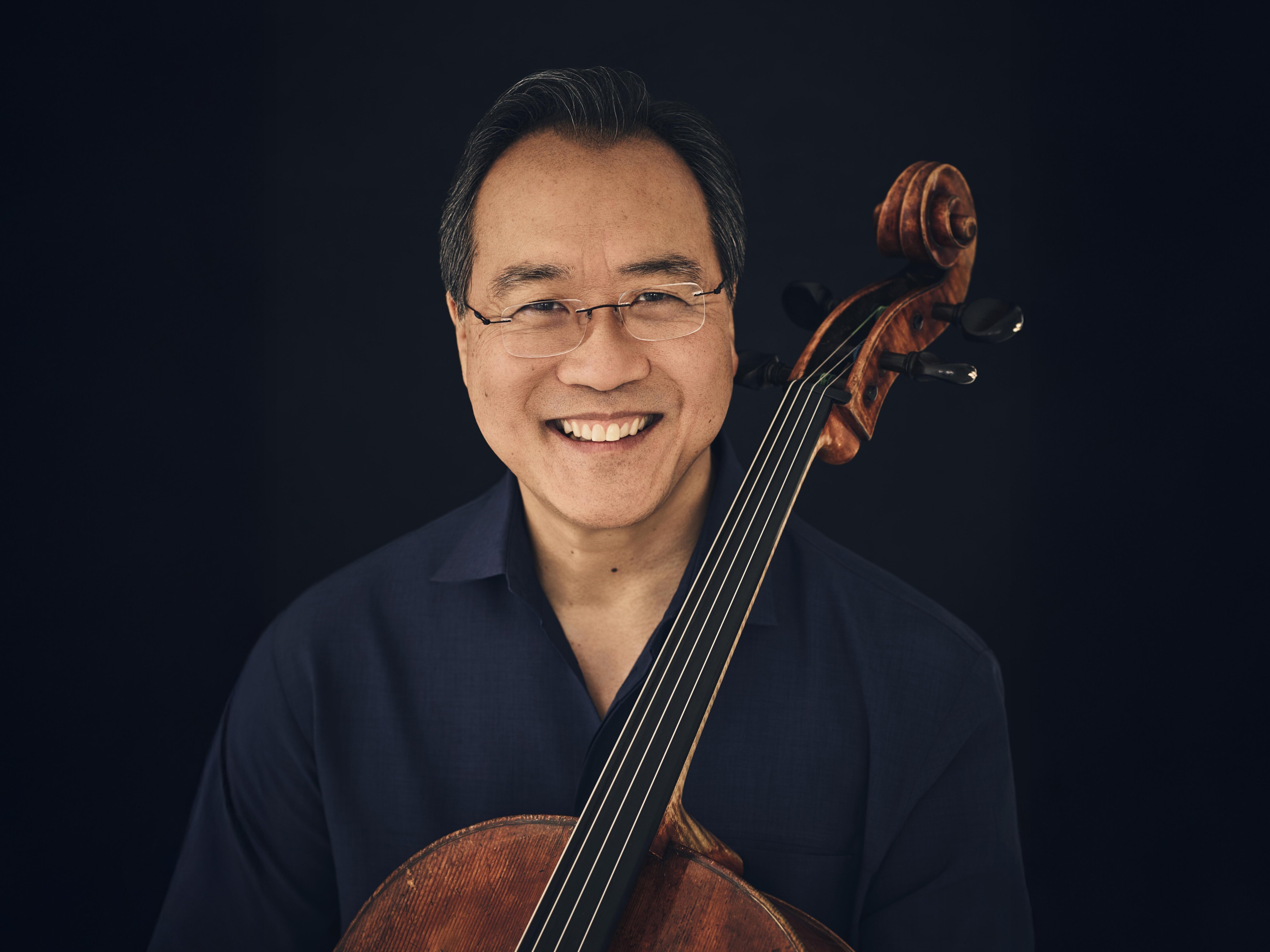Ein lächelnder Mann mit Brille und dunklem Haar hält vor einem dunklen Hintergrund ein Cello aufrecht vor sich und trägt ein schwarzes Hemd.