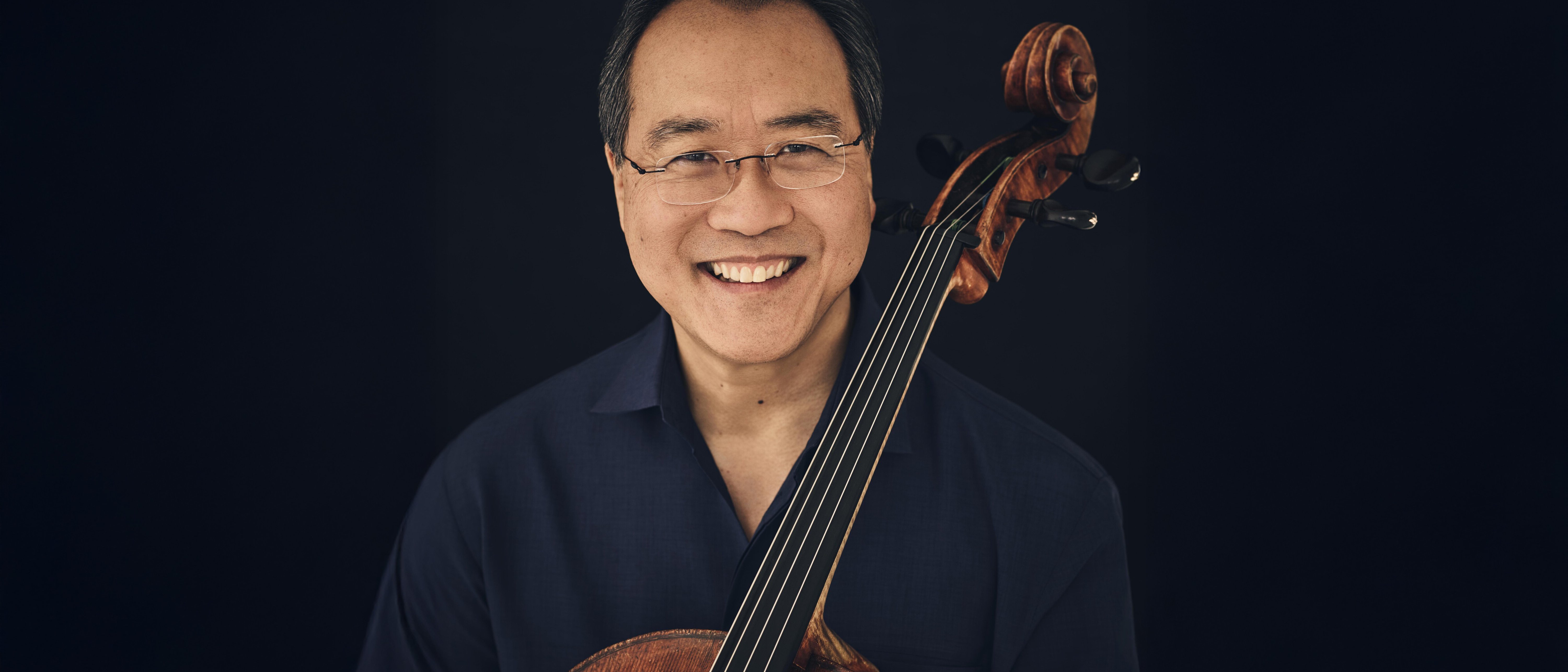 Ein lächelnder Mann mit Brille und dunklem Haar hält vor einem dunklen Hintergrund ein Cello aufrecht vor sich und trägt ein schwarzes Hemd.