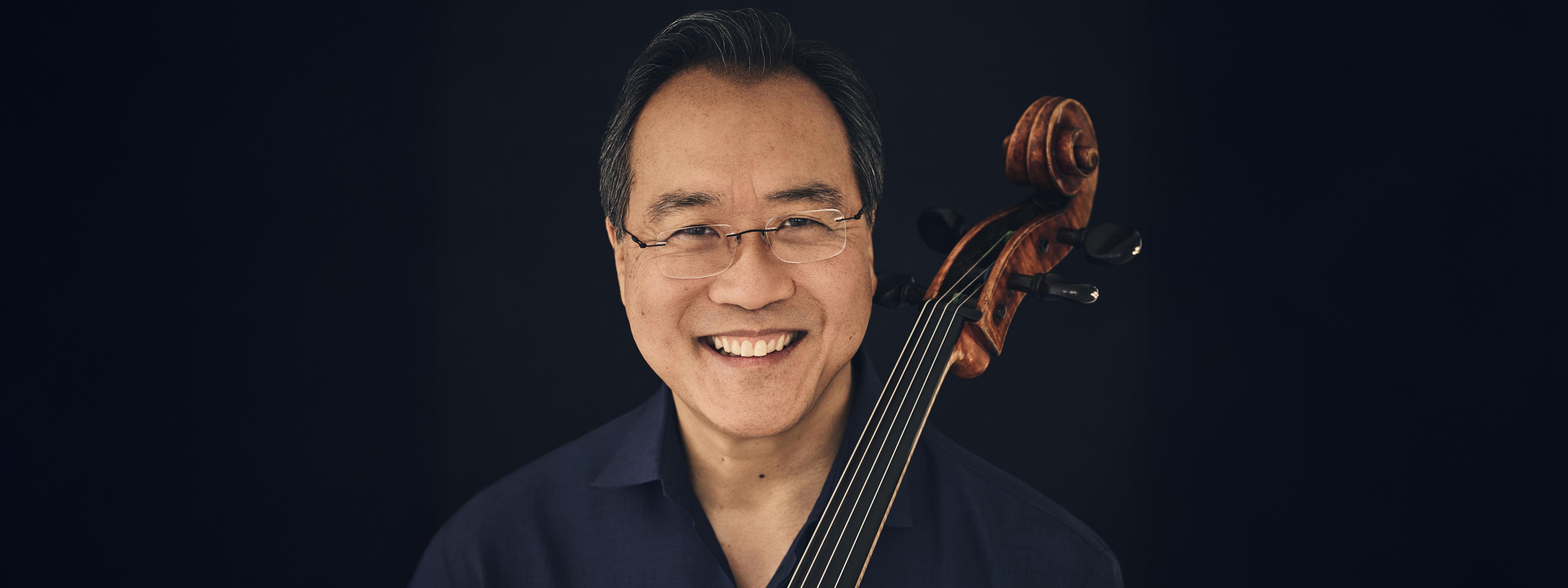 A smiling man with glasses holds a cello upright. He wears a black shirt and stands against a dark background, with the cello’s scroll visible near his shoulder.