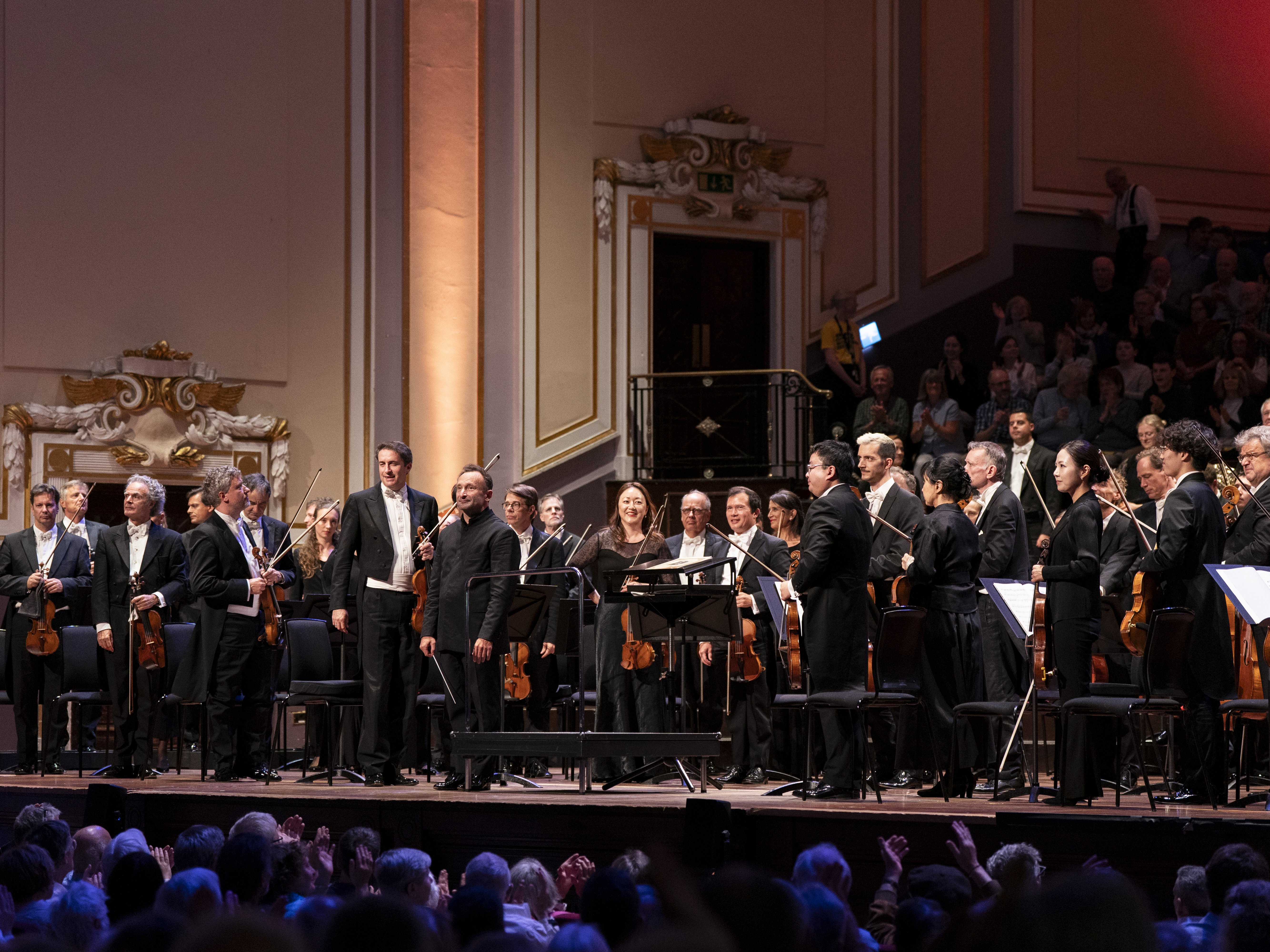 A symphony orchestra stands on stage in a grand concert hall, acknowledging applause from a large seated audience. Several musicians hold their instruments, and the conductor stands at the center.