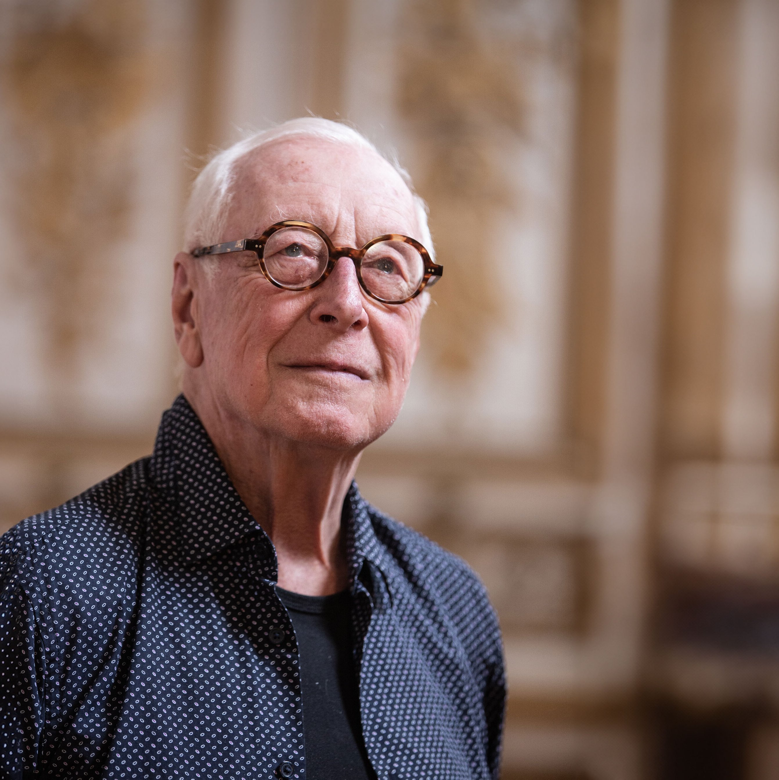 William Christie mit weißem Haar und runder Brille steht im Innenbereich. Er trägt ein dunkles, gemustertes Hemd über einem schwarzen Hemd. Der Hintergrund ist durch kunstvolle goldene und weiße Verzierungen verschwommen.