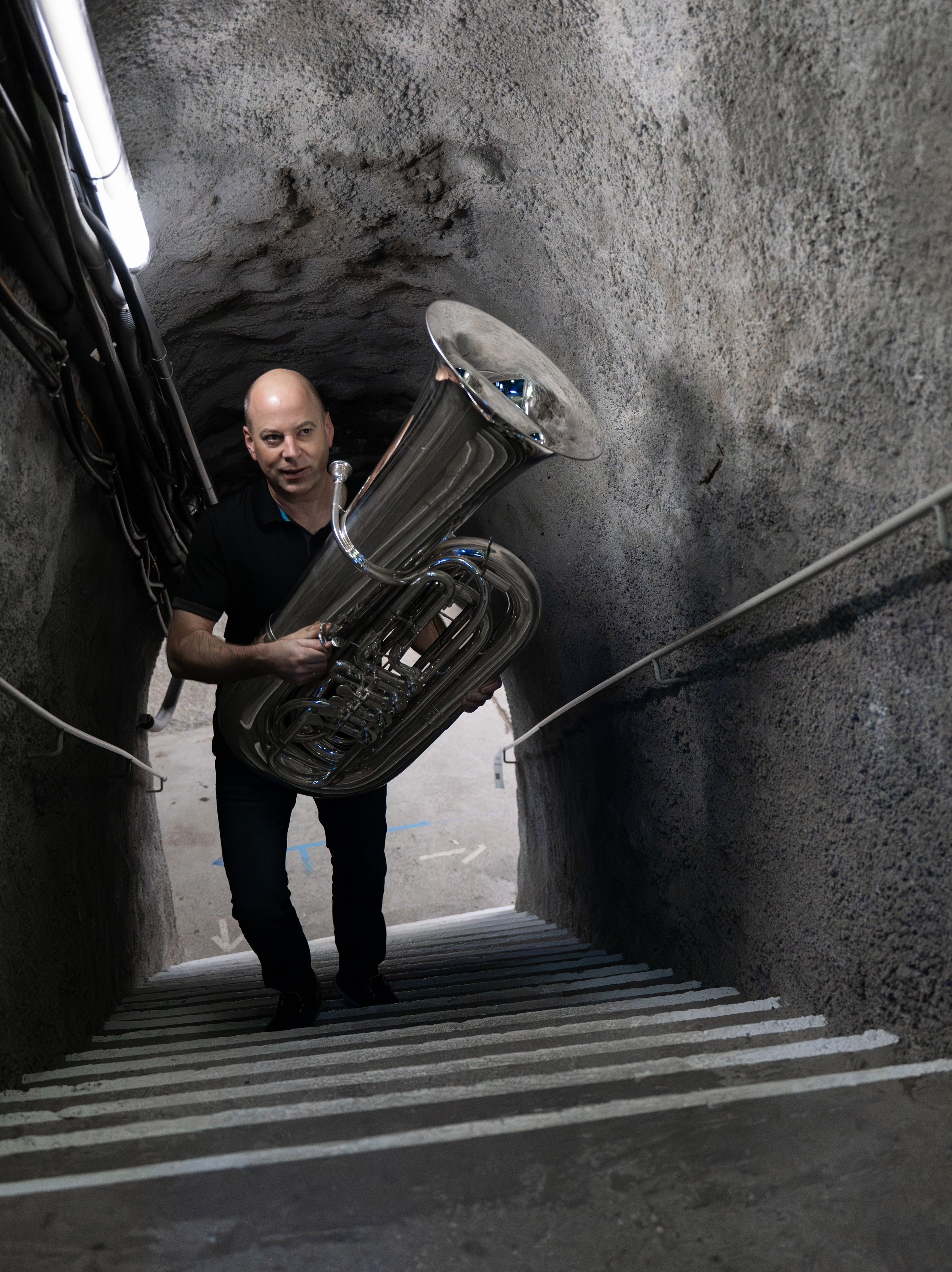 Ein Mann in dunkler Kleidung trägt eine große silberne Tuba eine schmale, schwach beleuchtete Betontreppe mit rauen Steinwänden und einem Metallgeländer hinauf.