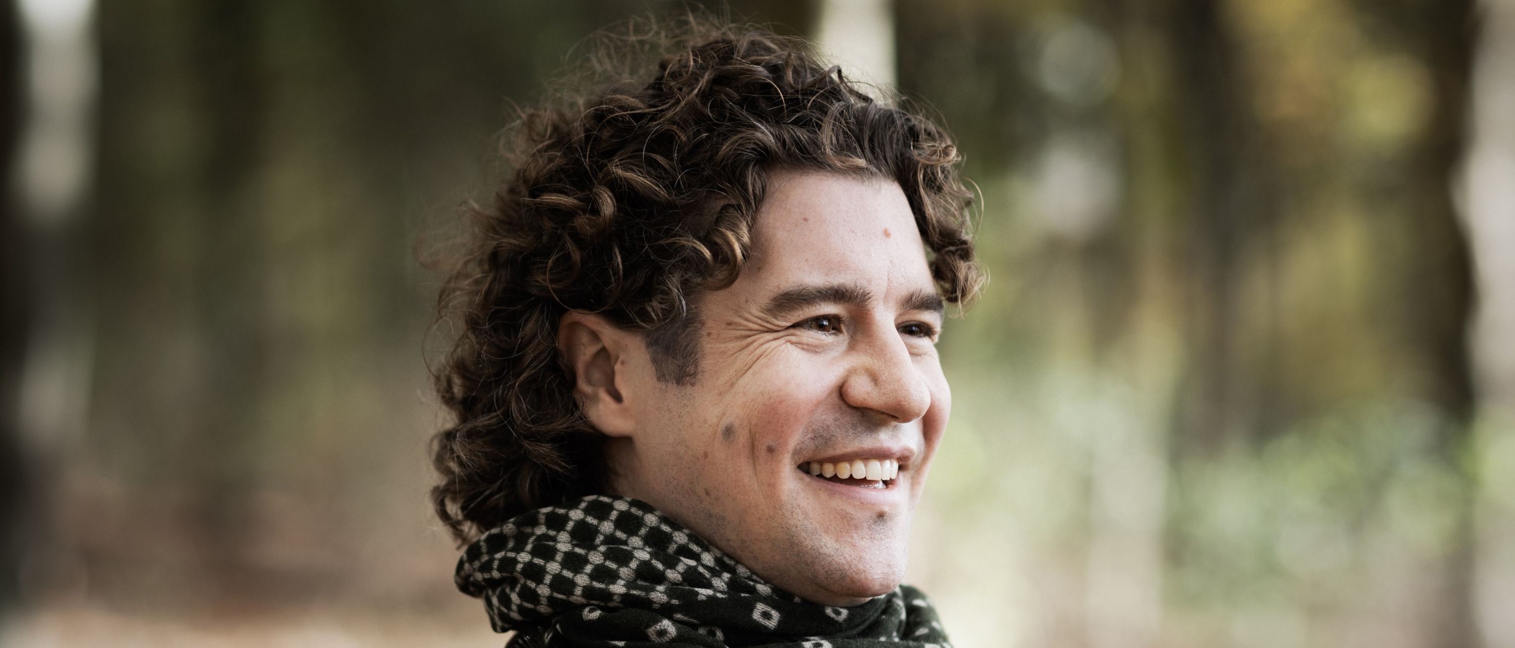 A man with curly hair smiles outdoors, wearing a dark jacket and a black-and-white patterned scarf. The background is blurred trees and foliage in a natural setting.