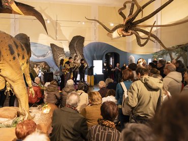 A crowd watches a small brass ensemble perform in a museum, surrounded by large prehistoric animal exhibits, including a dinosaur and a giant squid model hanging from the ceiling.