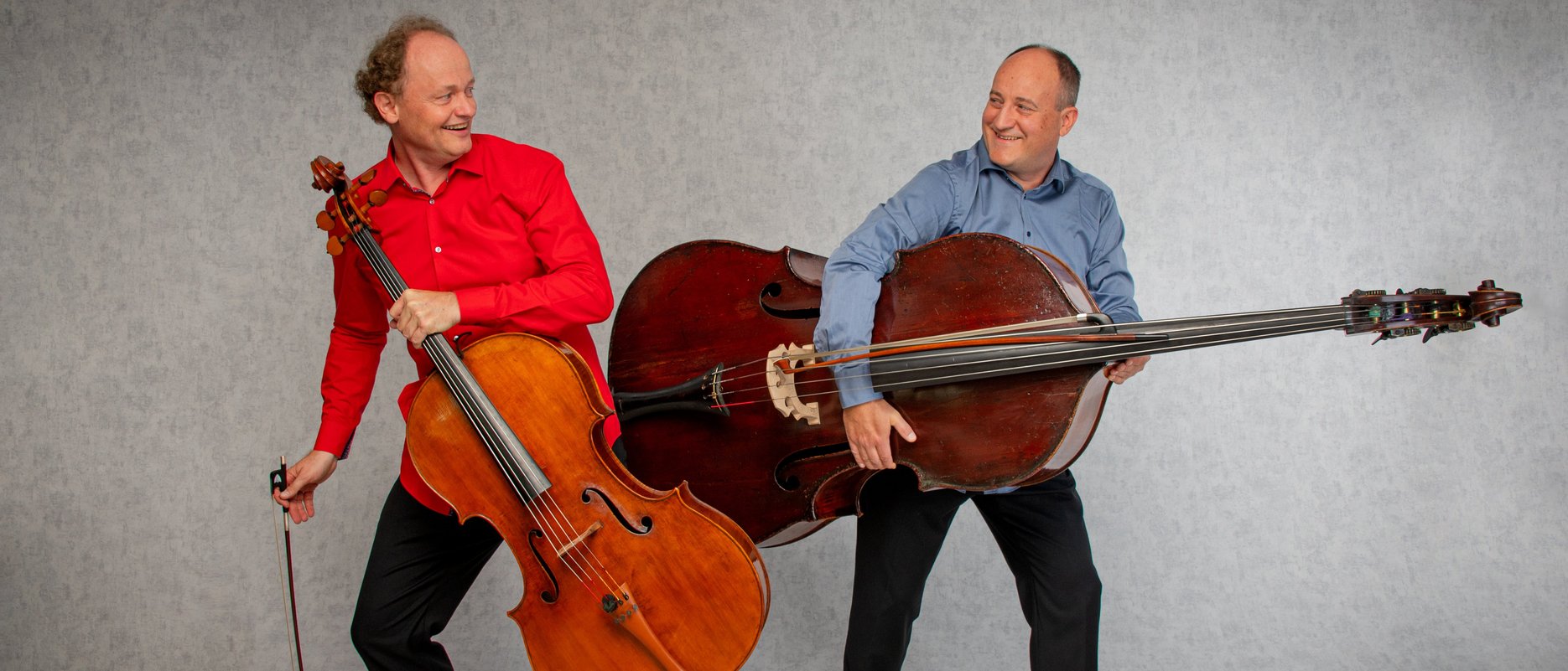 David Riniker and Janusz Widzyk stand back-to-back, holding large string instruments like guitars. One wears a red shirt and holds a cello, while the other in blue holds a double bass. Both appear playful and joyful.