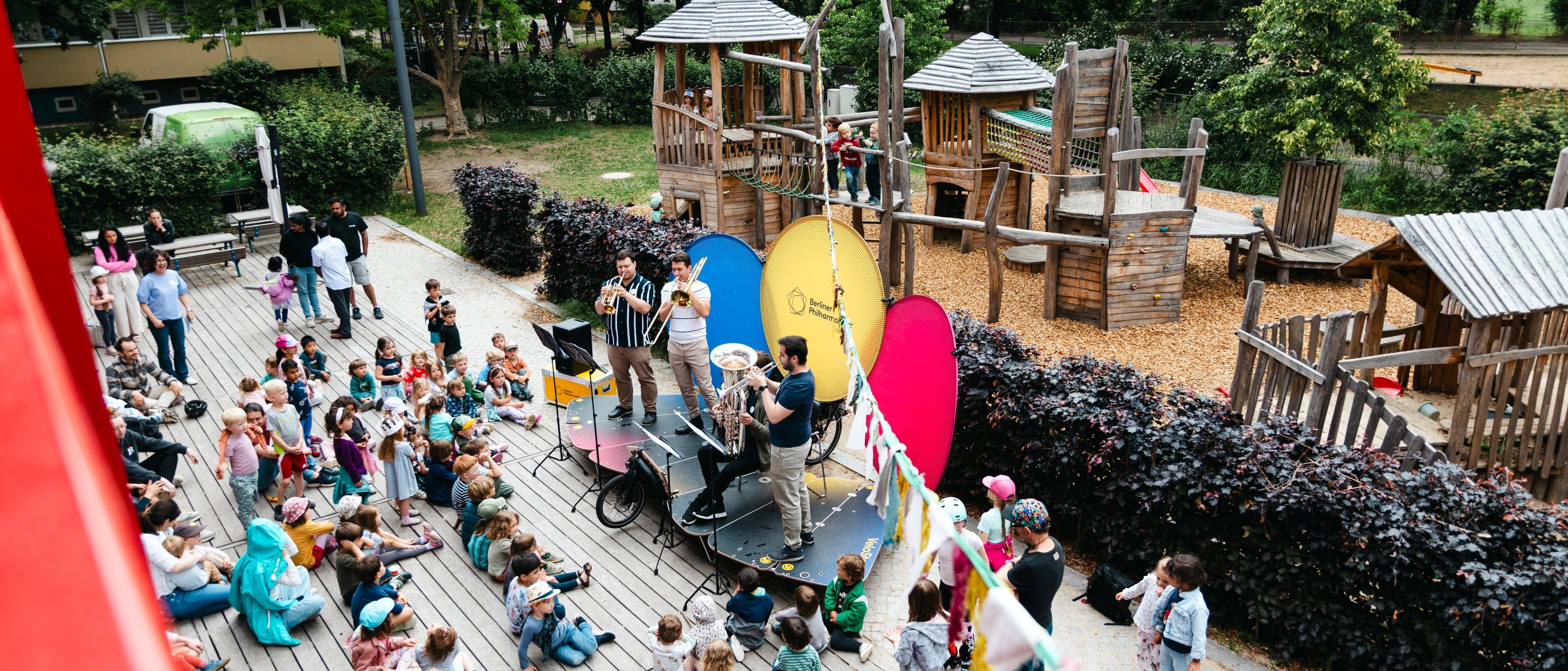 Ein Blechbläserensemble spielt auf einer Bühne vor einem Publikum aus Kindern und Erwachsenen auf einem Spielplatz im Freien, mit hölzernen Spielgeräten und Bäumen im Hintergrund.