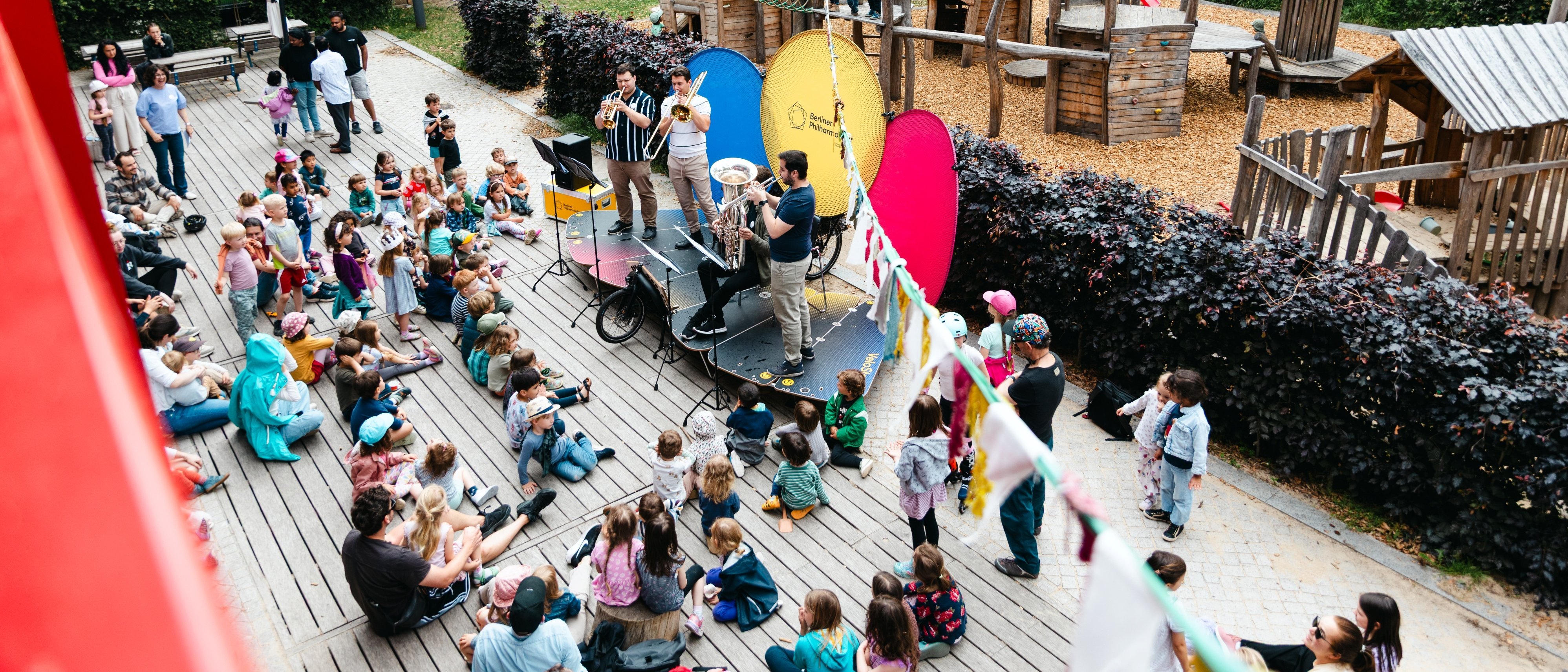 Ein Blechbläserensemble spielt auf einer Bühne vor einem Publikum aus Kindern und Erwachsenen auf einem Spielplatz im Freien, mit hölzernen Spielgeräten und Bäumen im Hintergrund.