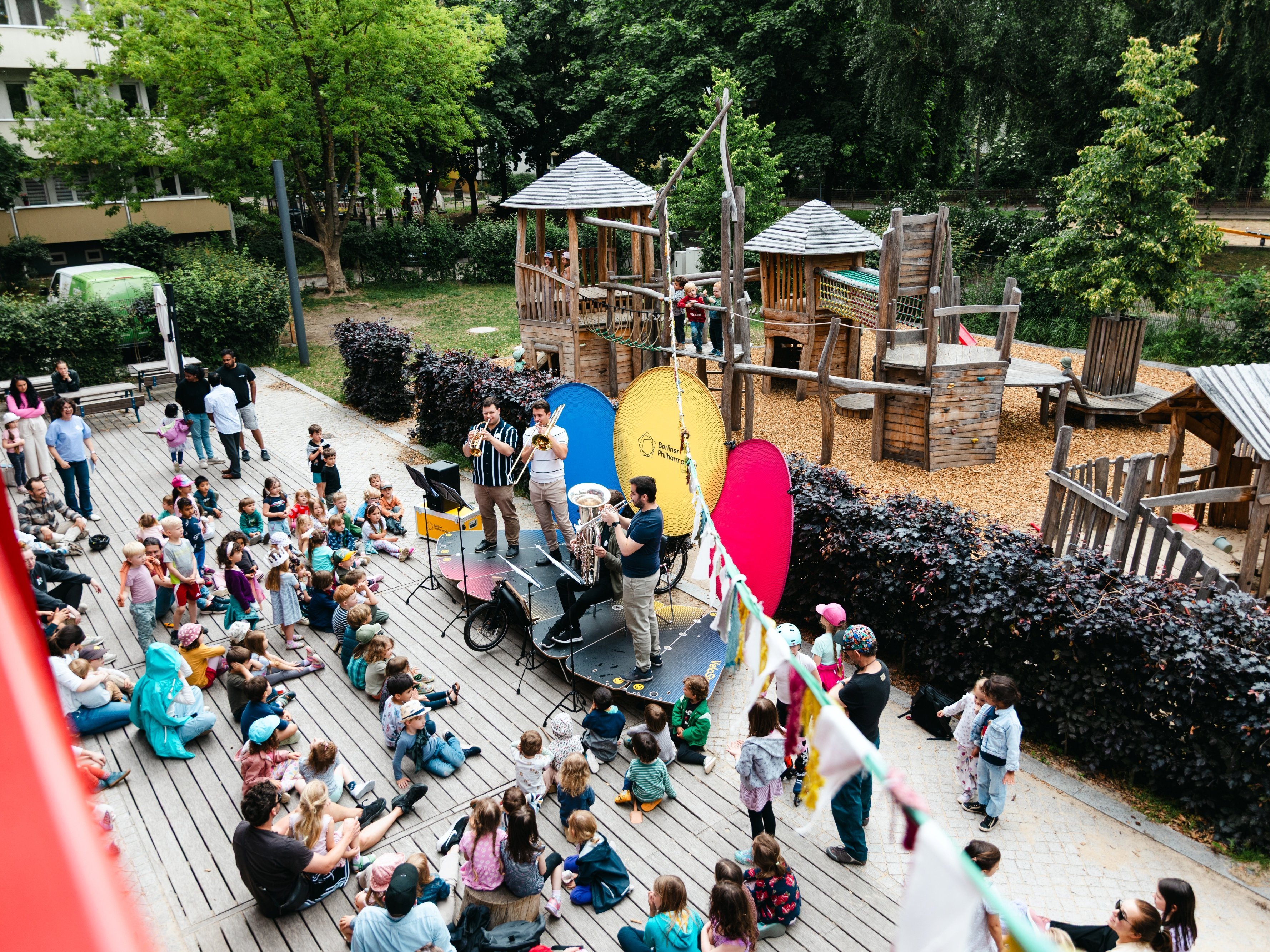 Ein Blechbläserensemble spielt auf einer Bühne vor einem Publikum aus Kindern und Erwachsenen auf einem Spielplatz im Freien, mit hölzernen Spielgeräten und Bäumen im Hintergrund.