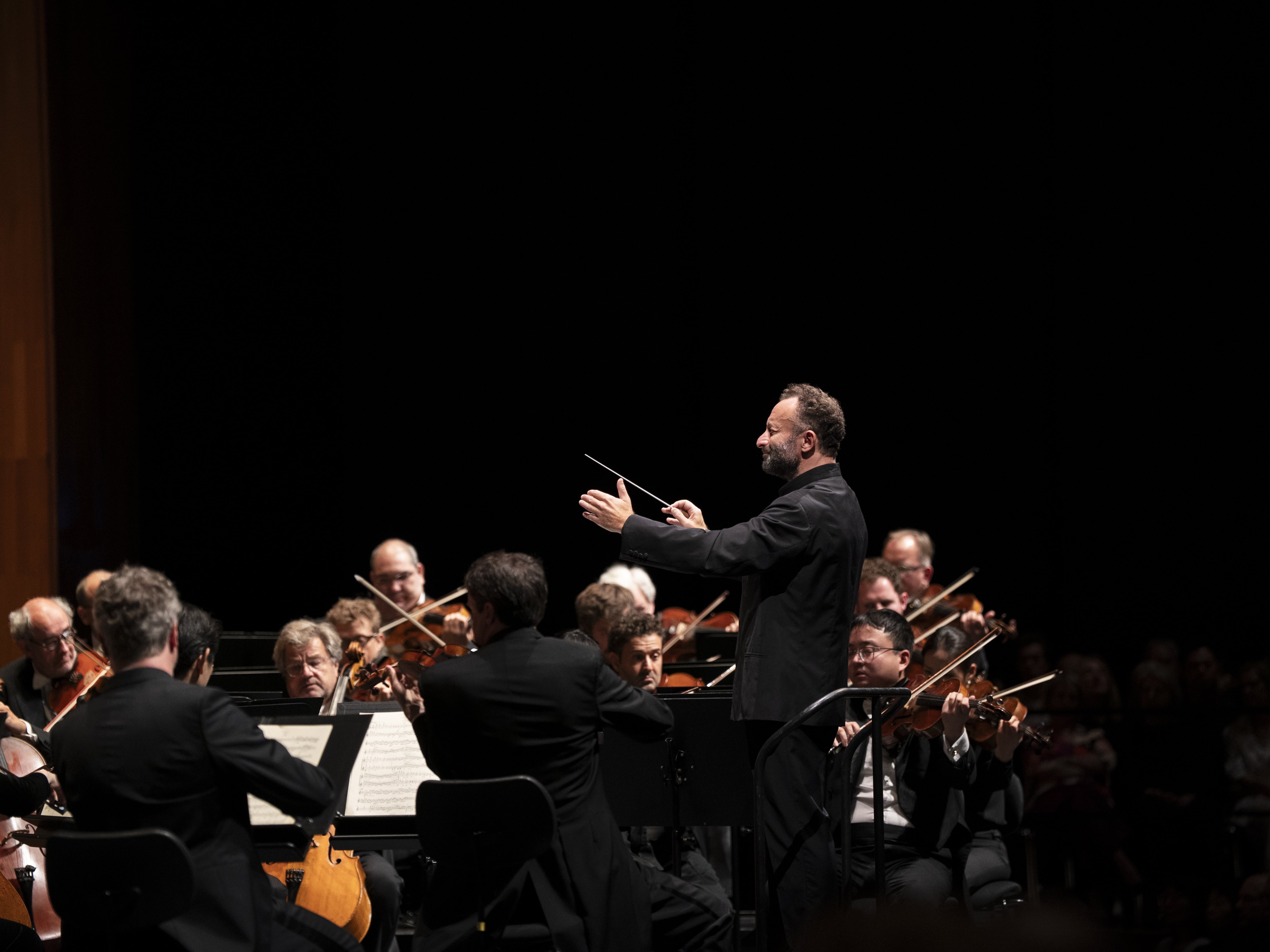 Ein Dirigent leitet ein Orchester während eines Konzerts. Die Musiker spielen Streichinstrumente und lesen dabei aus den Noten. Die Bühne ist schwach beleuchtet, sodass der Dirigent und die Musiker im Vordergrund besonders hervorstechen.