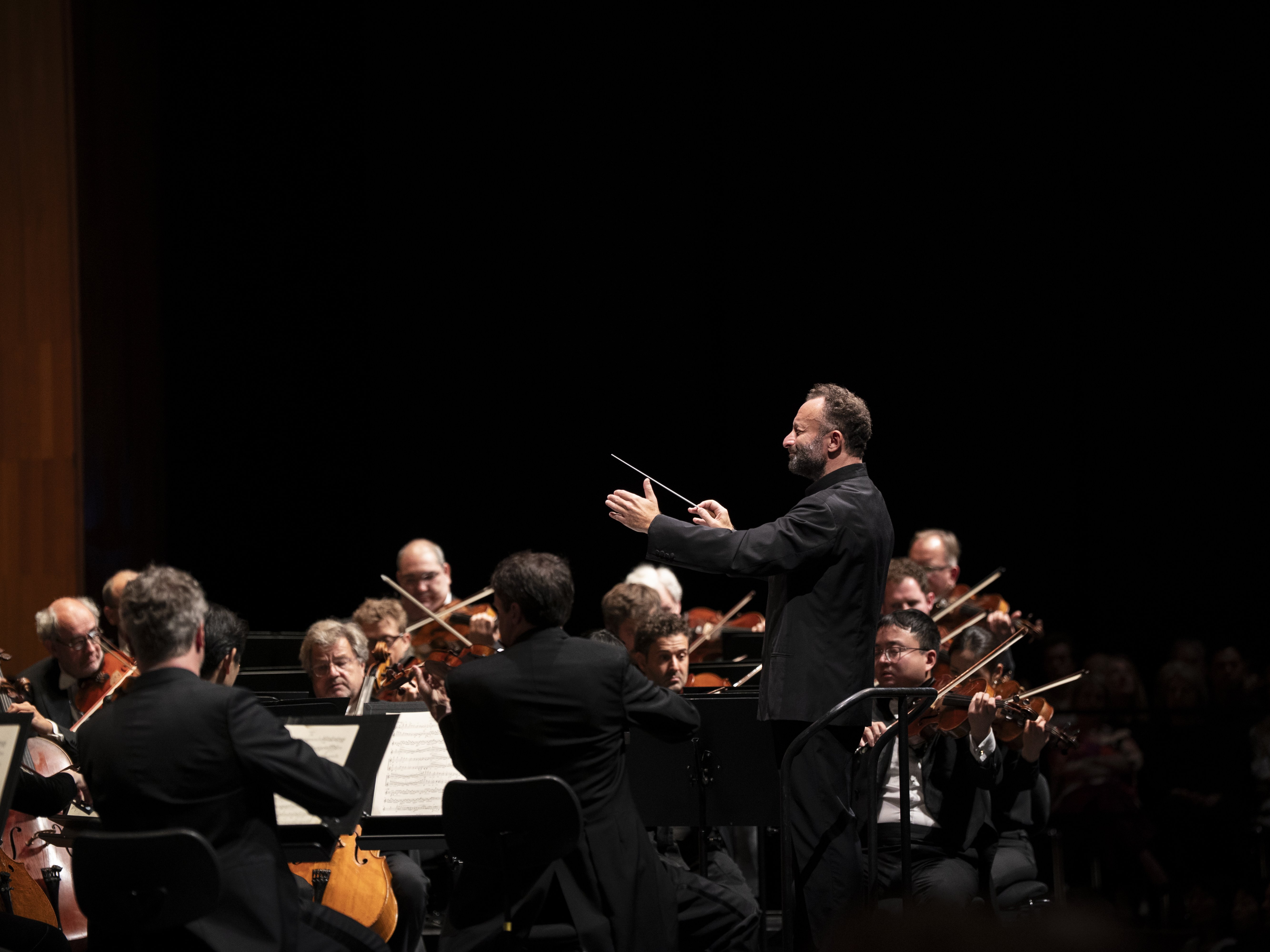 Ein Dirigent leitet ein Orchester während eines Konzerts. Die Musiker spielen Streichinstrumente und lesen dabei aus den Noten. Die Bühne ist schwach beleuchtet, sodass der Dirigent und die Musiker im Vordergrund besonders hervorstechen.