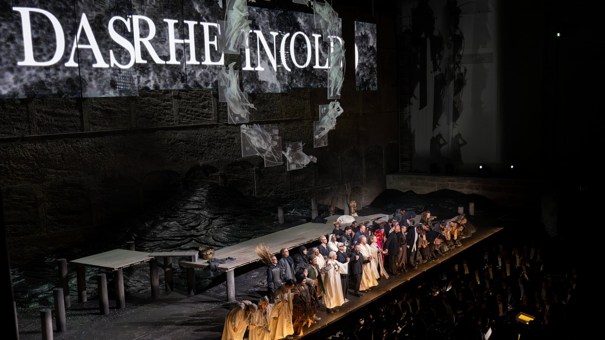 Kirill Petrenko sowie eine große Gruppe von Darstellern in Kostümen stehen auf der Bühne, hält sich an den Händen und verbeugen sich vor dem applaudierenden Publikum nach einer Theateraufführung, während im Hintergrund Kulissen und dramatische Beleuchtung zu sehen sind.