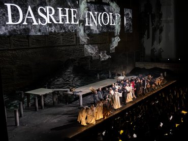 Kirill Petrenko sowie eine große Gruppe von Darstellern in Kostümen stehen auf der Bühne, hält sich an den Händen und verbeugen sich vor dem applaudierenden Publikum nach einer Theateraufführung, während im Hintergrund Kulissen und dramatische Beleuchtung zu sehen sind.