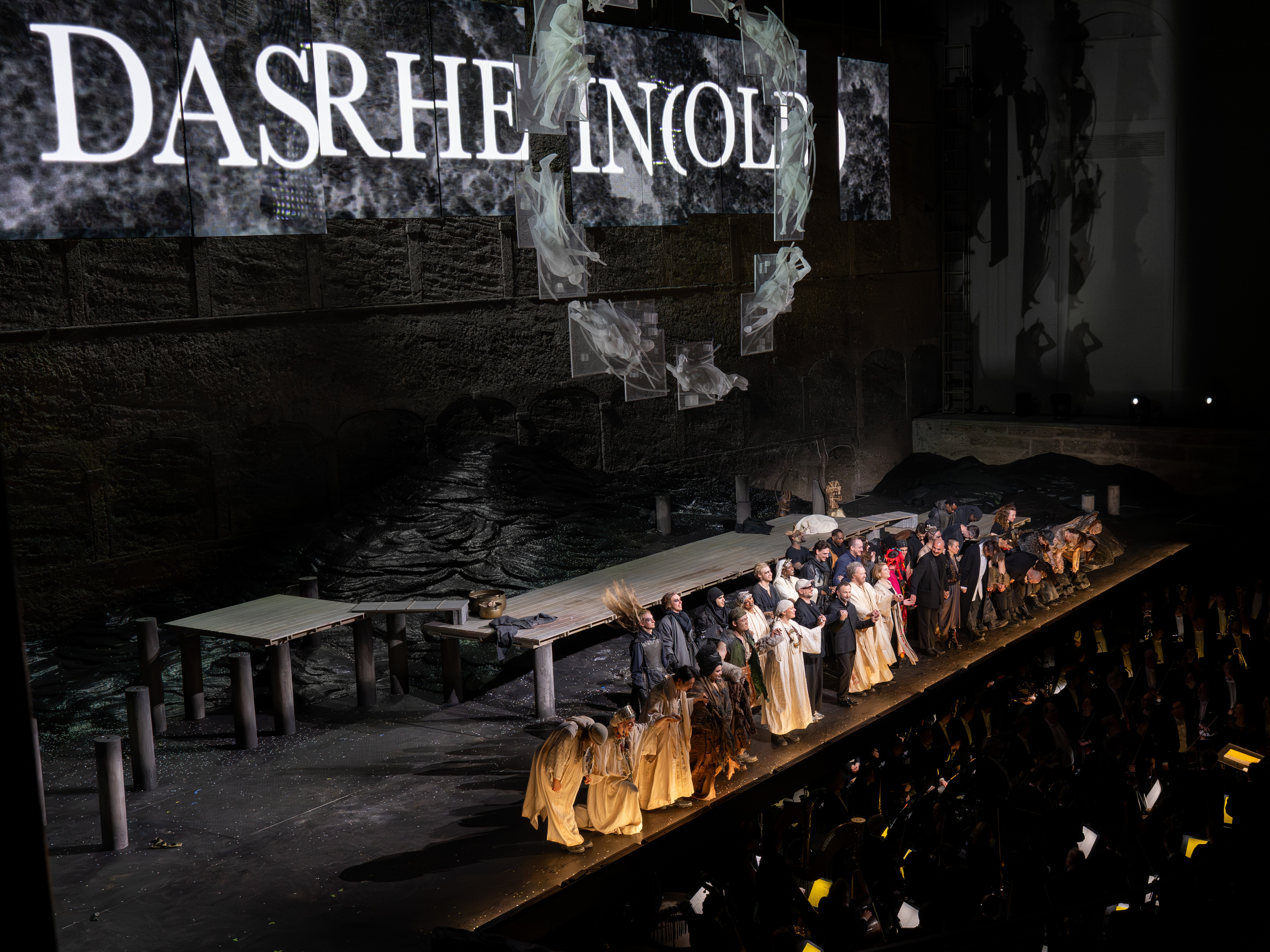Kirill Petrenko sowie eine große Gruppe von Darstellern in Kostümen stehen auf der Bühne, hält sich an den Händen und verbeugen sich vor dem applaudierenden Publikum nach einer Theateraufführung, während im Hintergrund Kulissen und dramatische Beleuchtung zu sehen sind.