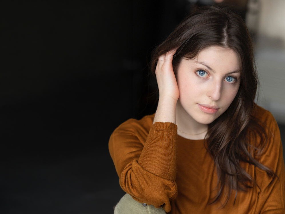 Eine junge Frau mit langen braunen Haaren und blauen Augen, die einen braunen Pullover trägt, sitzt mit einer Hand an ihrem Kopf und blickt sanft in die Kamera vor einem dunklen, verschwommenen Hintergrund.