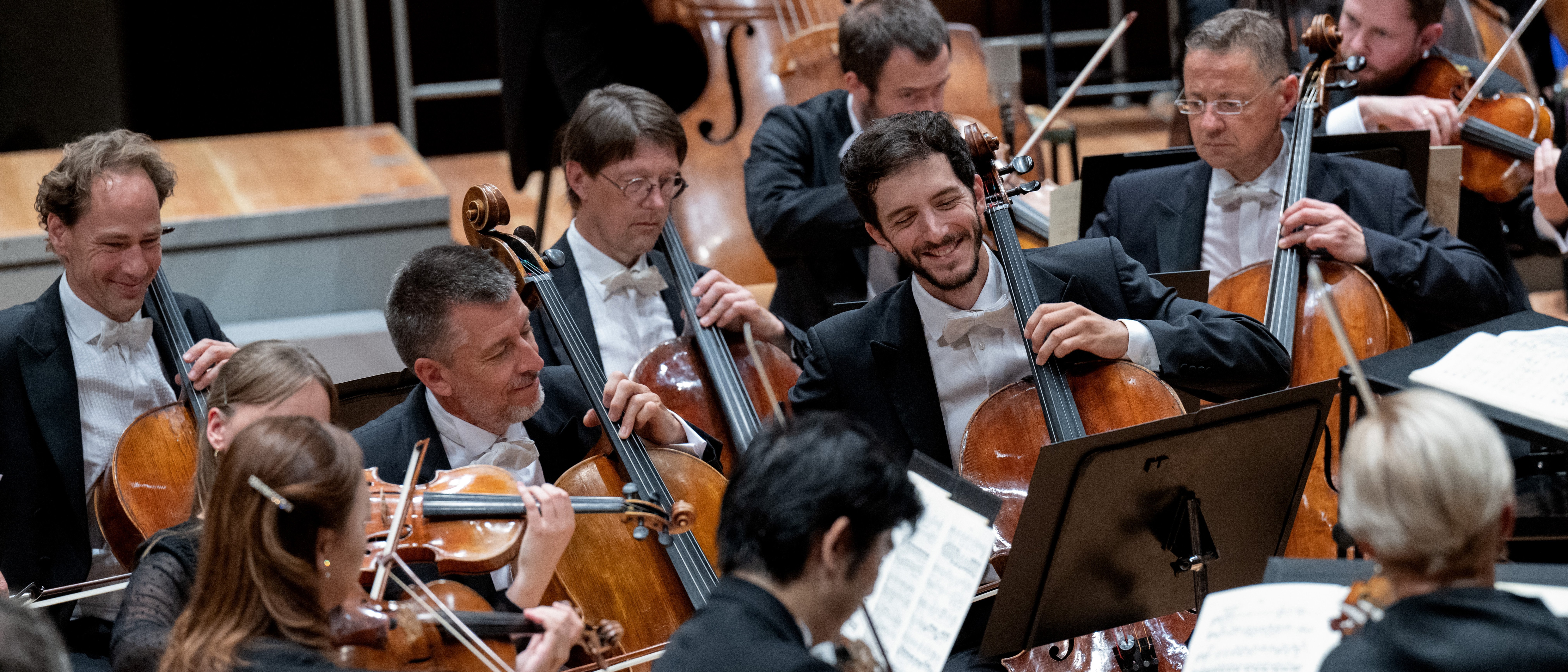 Mehrere Musikerinnen in formeller Kleidung spielen Celli und Geigen in einem Orchester. Sie sitzen eng beieinander, lesen Noten und einige lächeln, während sie auf der Bühne spielen.