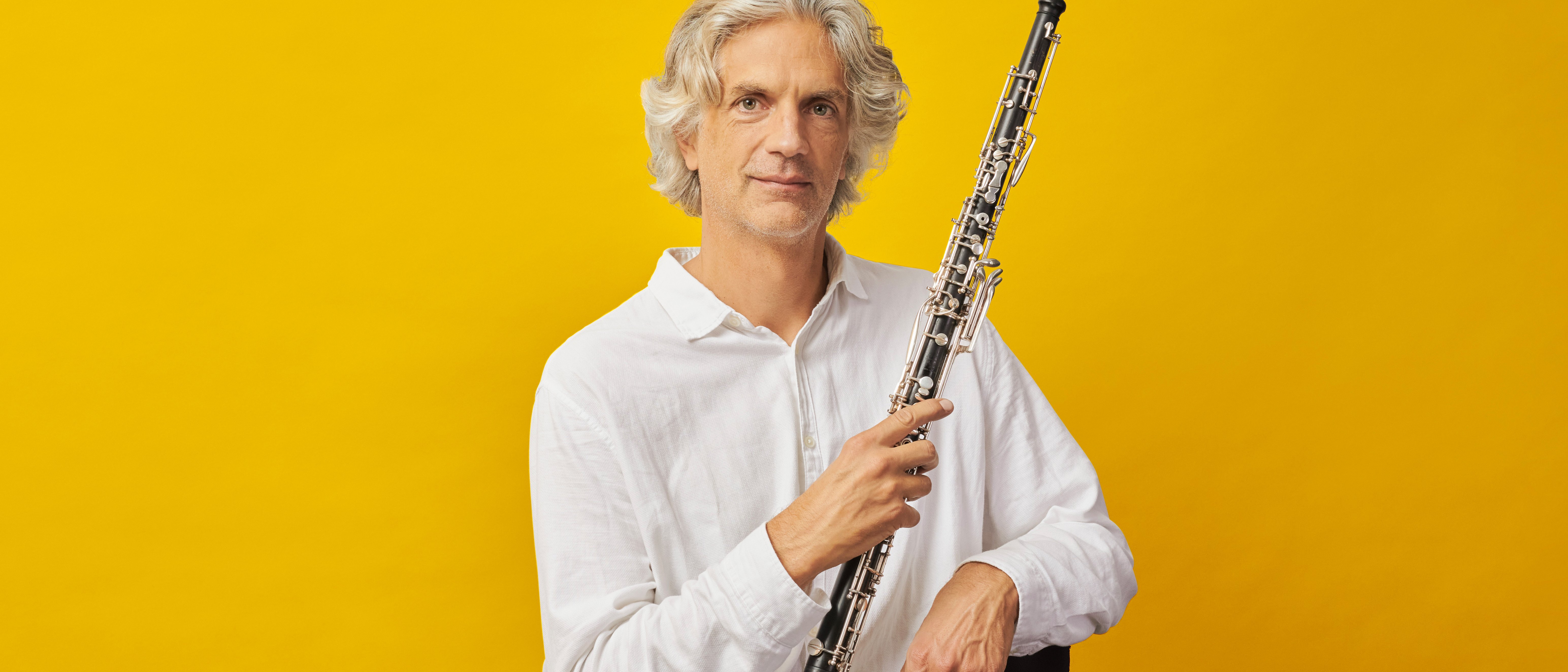Dominik Wollenweber mit grauem Haar und weißem Hemd sitzt auf einem Stuhl und hält eine Oboe aufrecht vor einem leuchtend gelben Hintergrund.