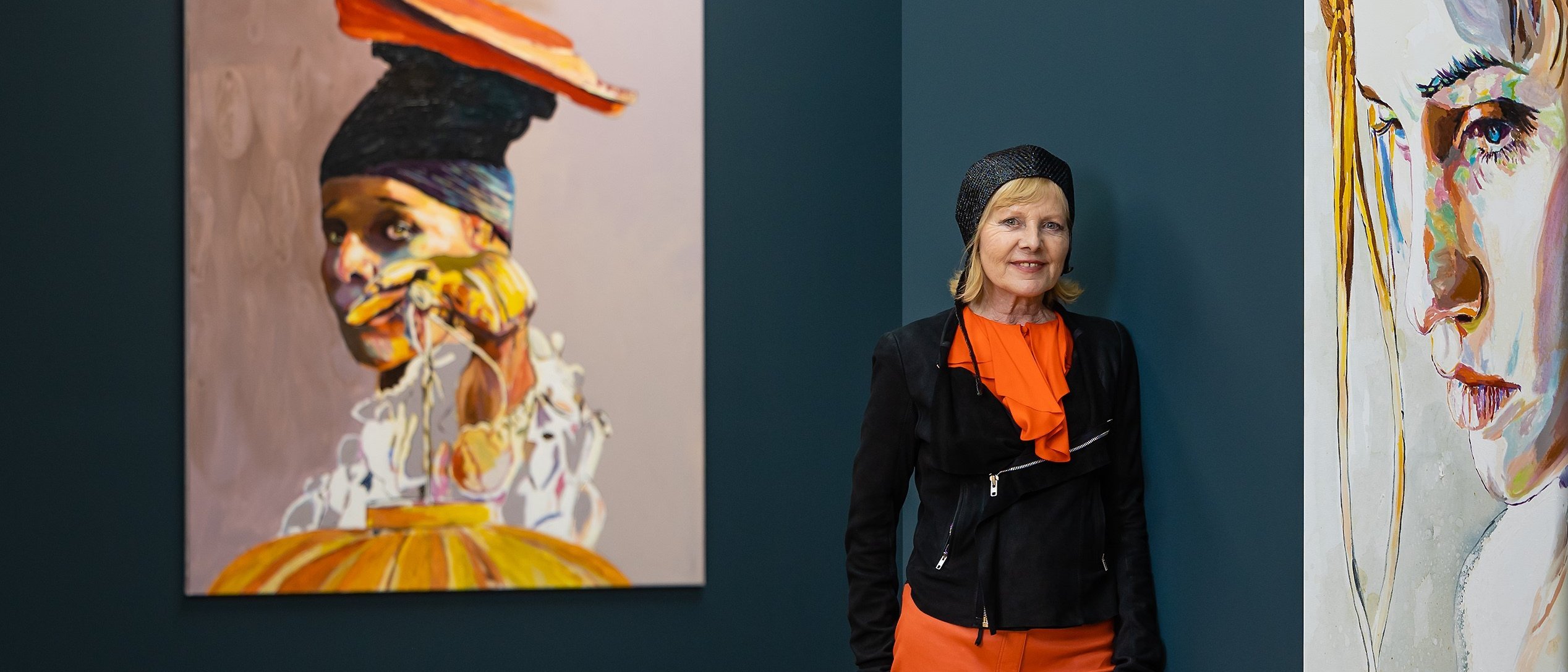 Cornelia Schleime, wearing orange trousers, a black jacket and yellow shoes, stands in an art gallery with a small dog on a leash, next to two large portrait paintings on dark blue walls.