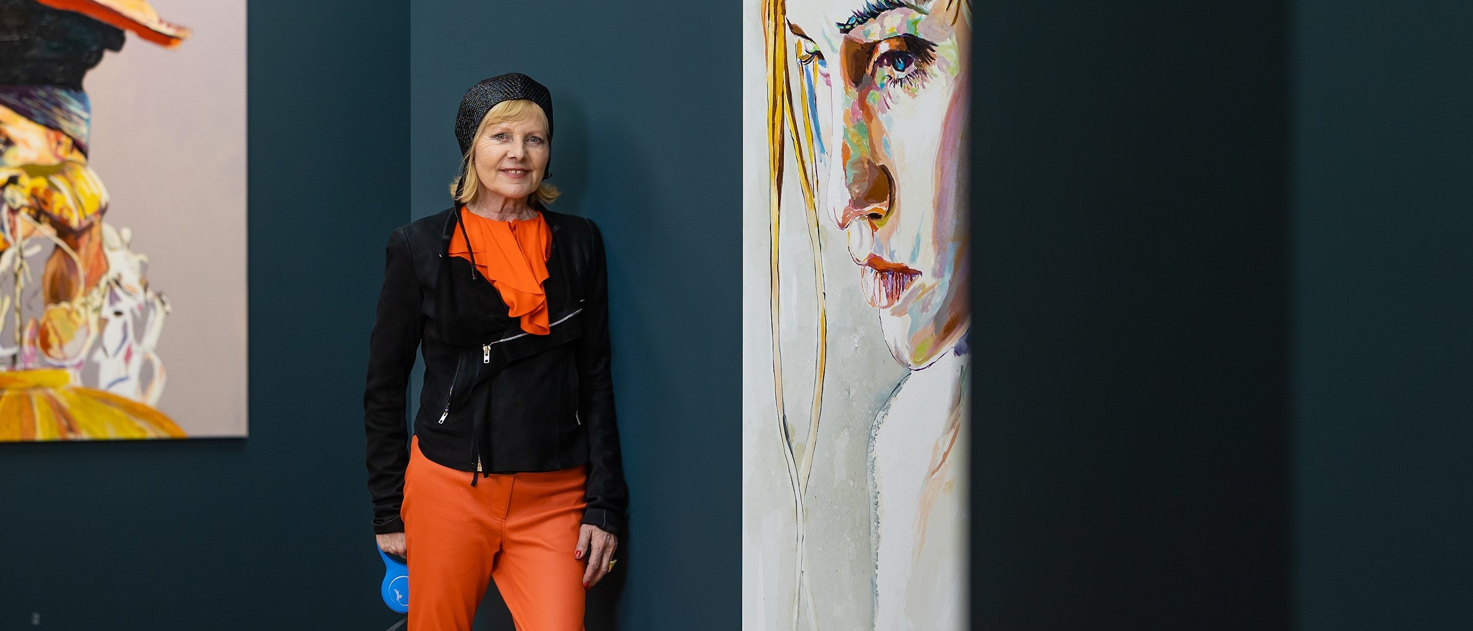 Cornelia Schleime in orangefarbener Hose, schwarzer Jacke und gelben Schuhen steht in einer Kunstgalerie mit einem kleinen Hund an der Leine, neben zwei großen Porträtgemälden an dunkelblauen Wänden.