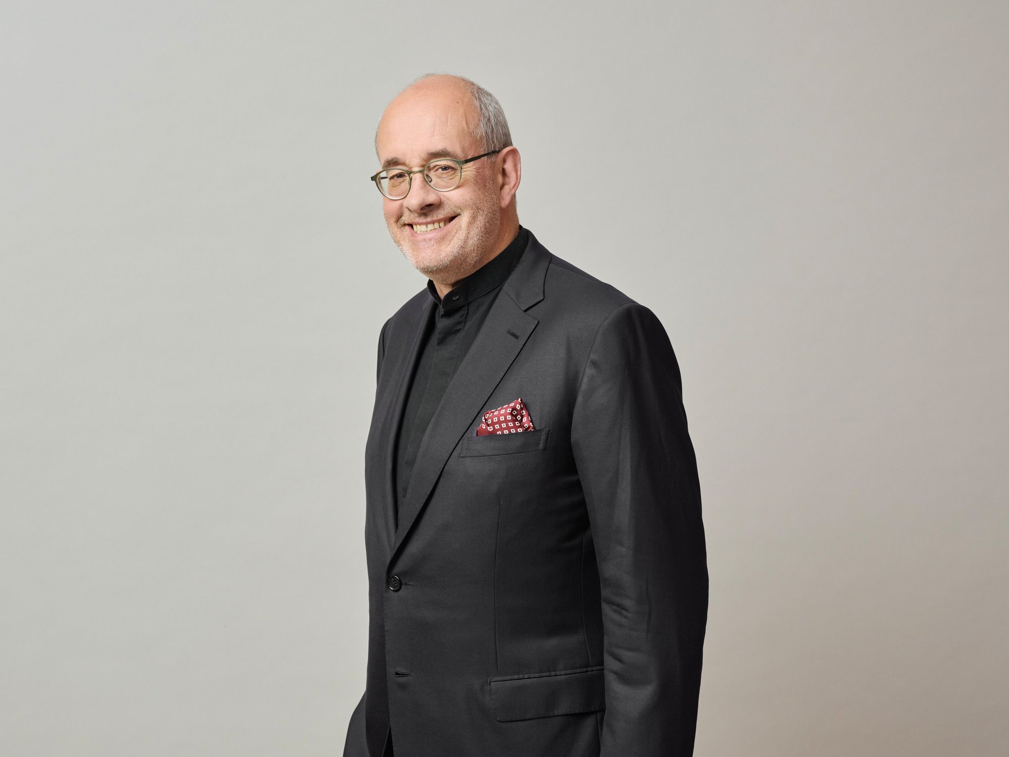 Ein älterer Mann mit Brille und Bart lächelt; er trägt einen dunklen Anzug, ein schwarzes Hemd und ein rot gemustertes Einstecktuch und steht vor einem schlichten hellgrauen Hintergrund.