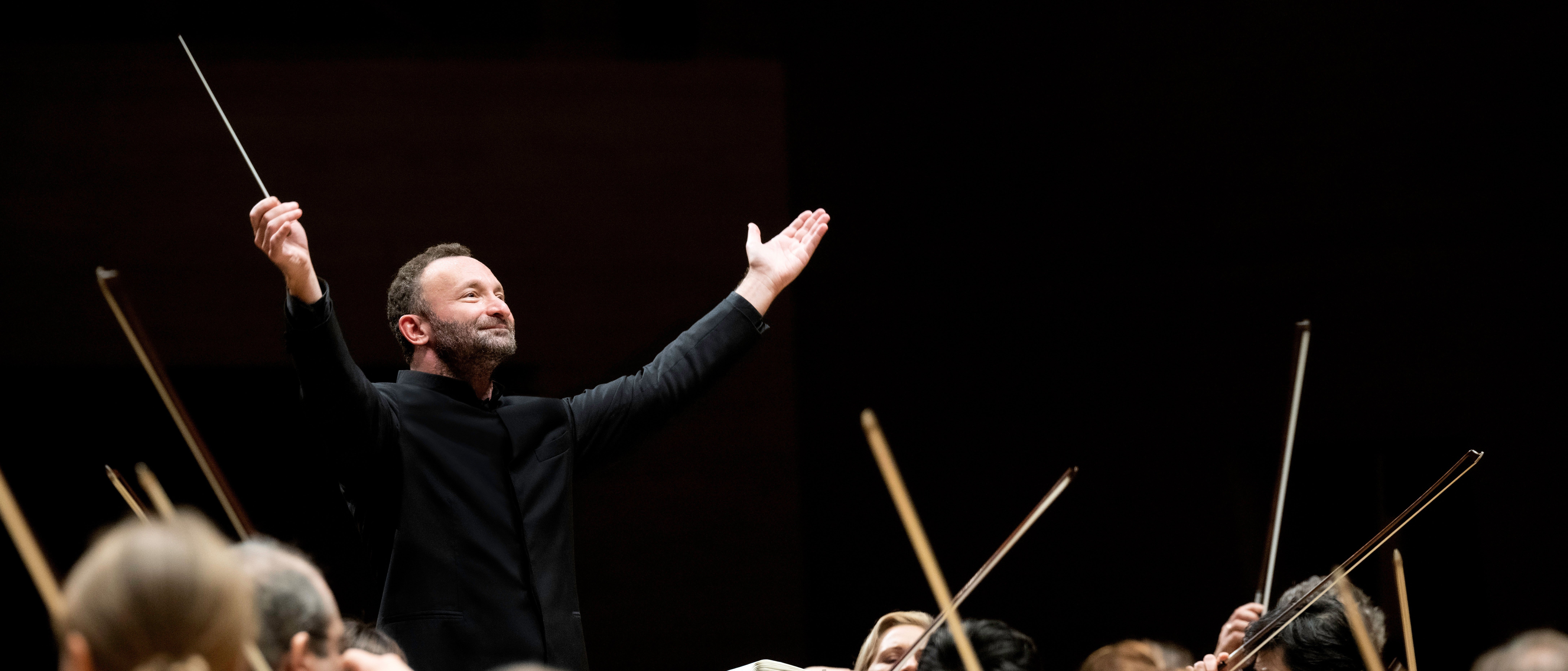 Kirill Petrenko steht vor dem Orchester und hebt beide Arme zu einer großen Geste nach oben. Er trägt einen schwarzen Anzug. Im Vordergrund sind Musiker von hinten zu sehen.