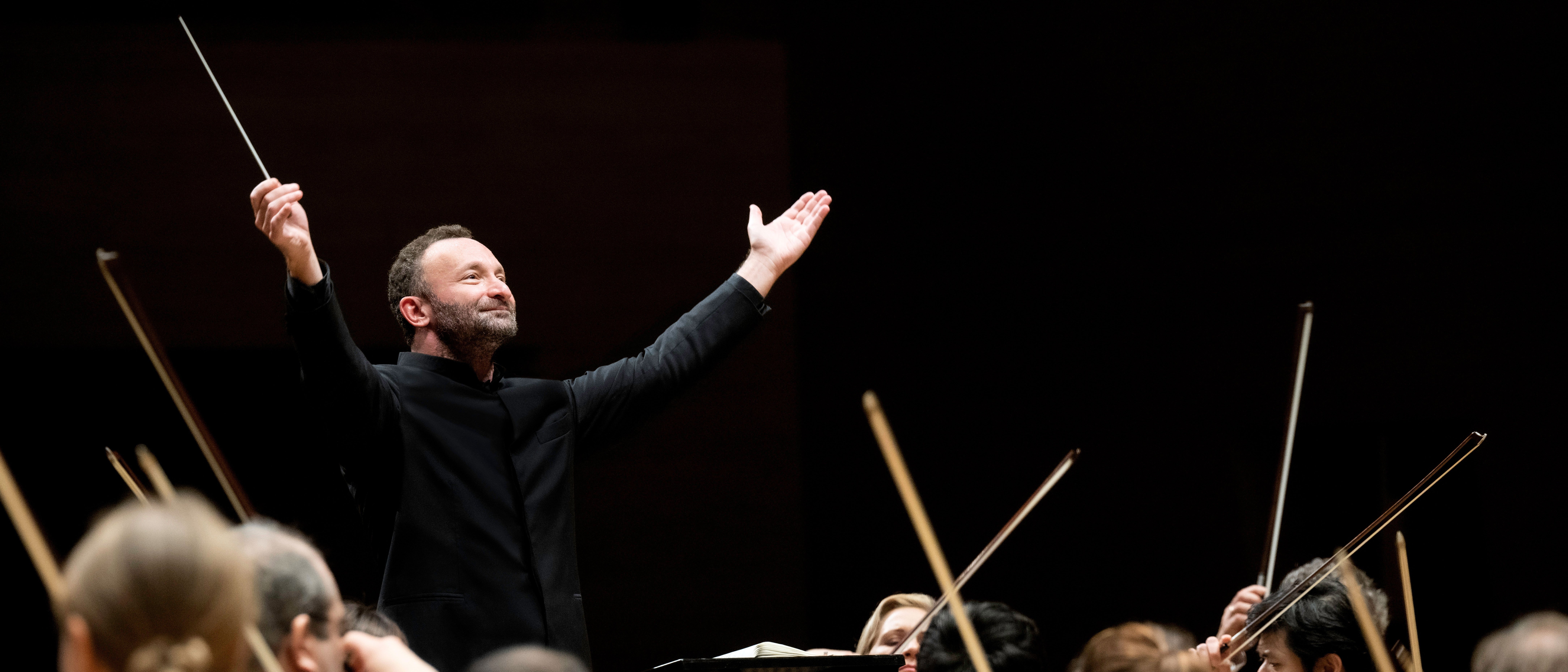 Kirill Petrenko steht vor dem Orchester und hebt beide Arme zu einer großen Geste nach oben. Er trägt einen schwarzen Anzug. Im Vordergrund sind Musiker von hinten zu sehen.