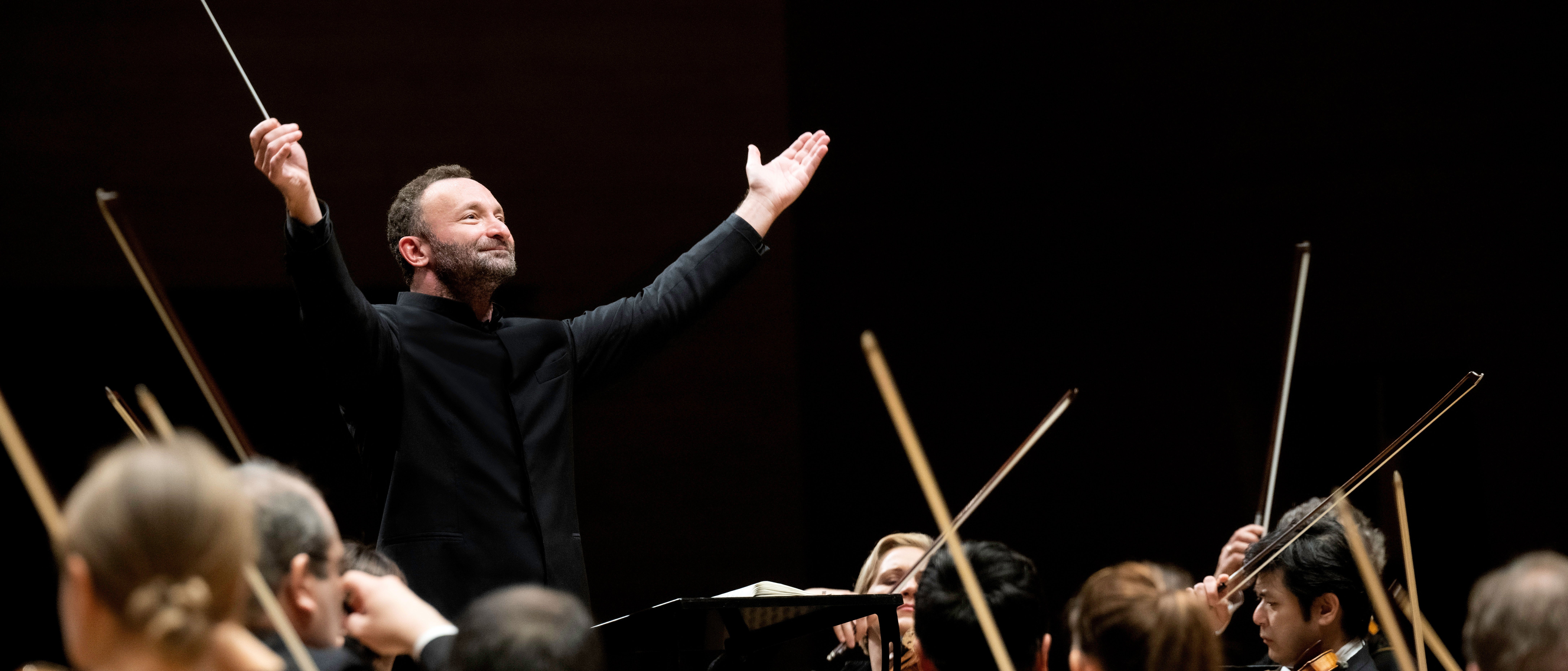 Kirill Petrenko stands in front of the orchestra and raises both arms upwards in a grand gesture. He is wearing a black suit. Musicians can be seen from behind in the foreground.