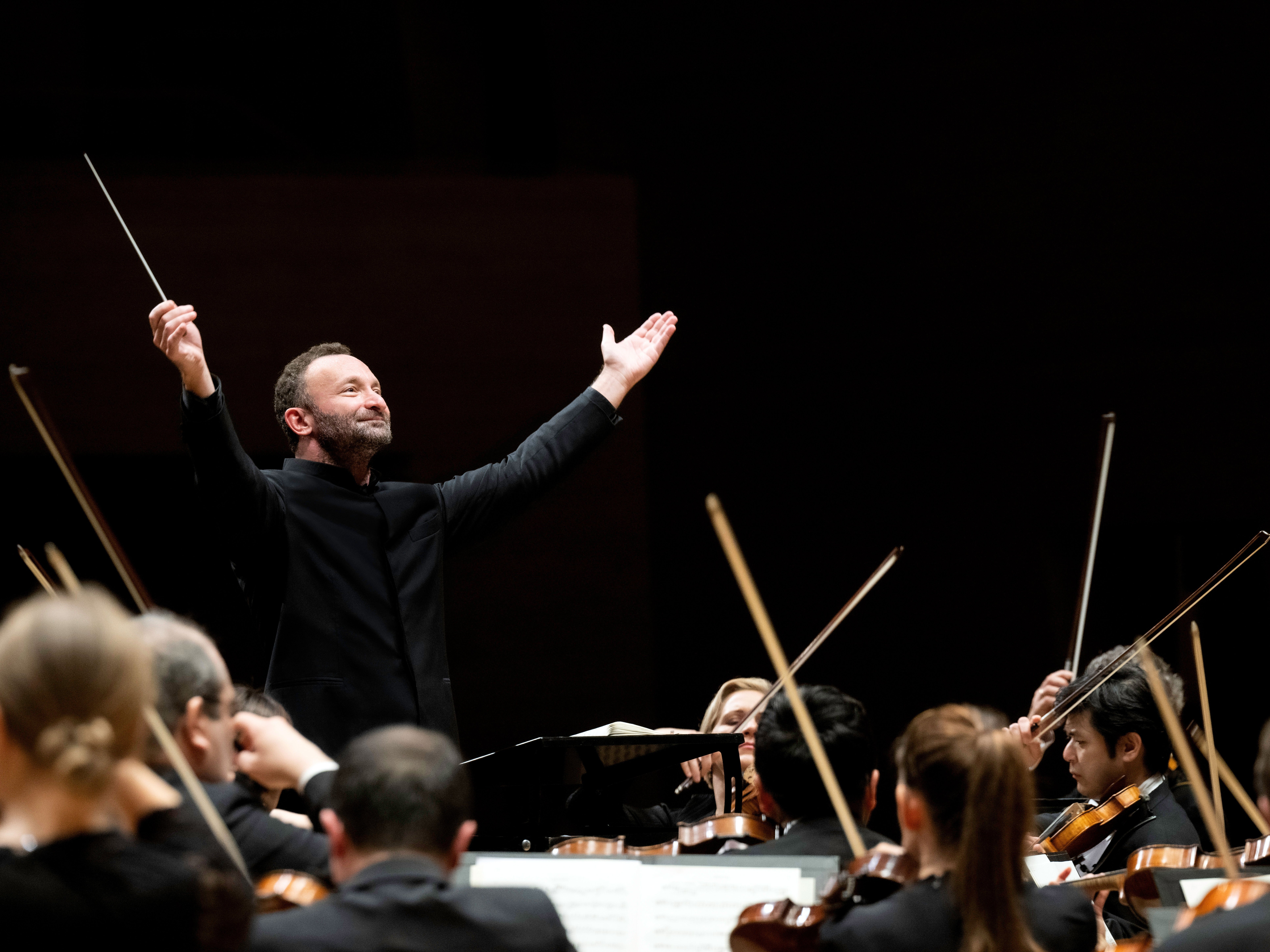Kirill Petrenko steht vor dem Orchester und hebt beide Arme zu einer großen Geste nach oben. Er trägt einen schwarzen Anzug. Im Vordergrund sind Musiker von hinten zu sehen.