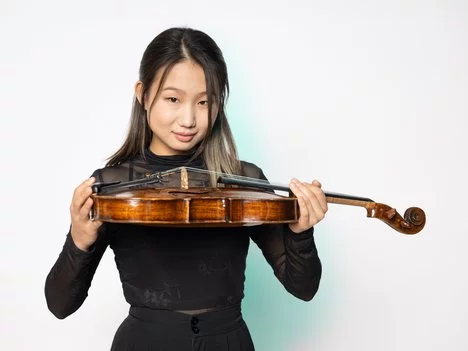 Jimin Jang in a black long-sleeve top holds a violin horizontally under her chin and smiles slightly, standing against a plain white background.