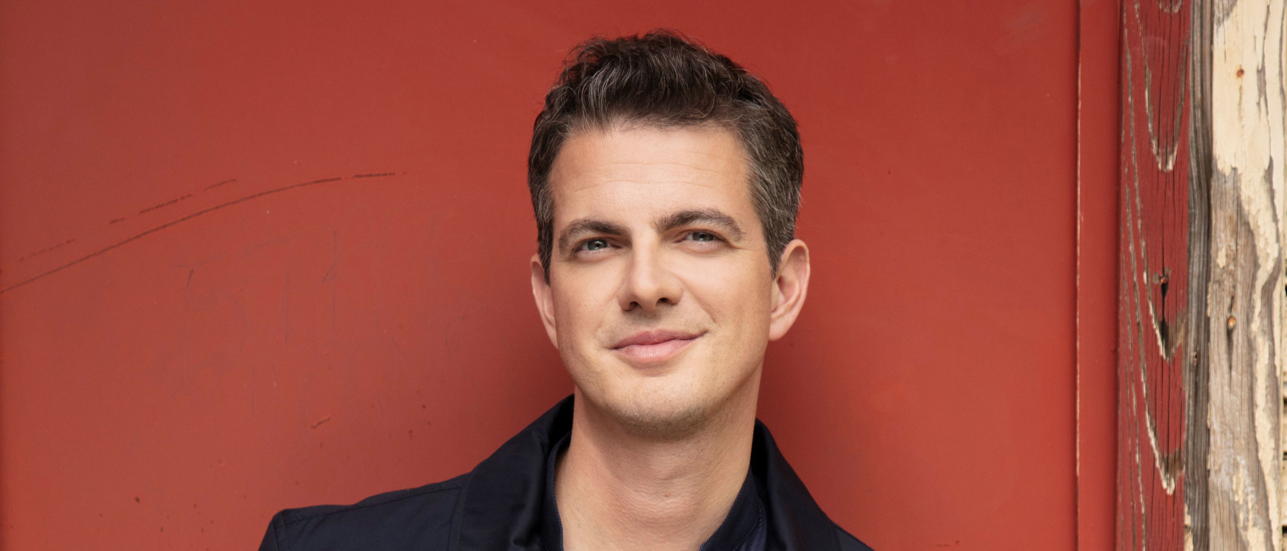 A man with short dark hair, wearing a navy jacket over a dark shirt, stands in front of a red wall with a worn wooden edge, smiling slightly at the camera.