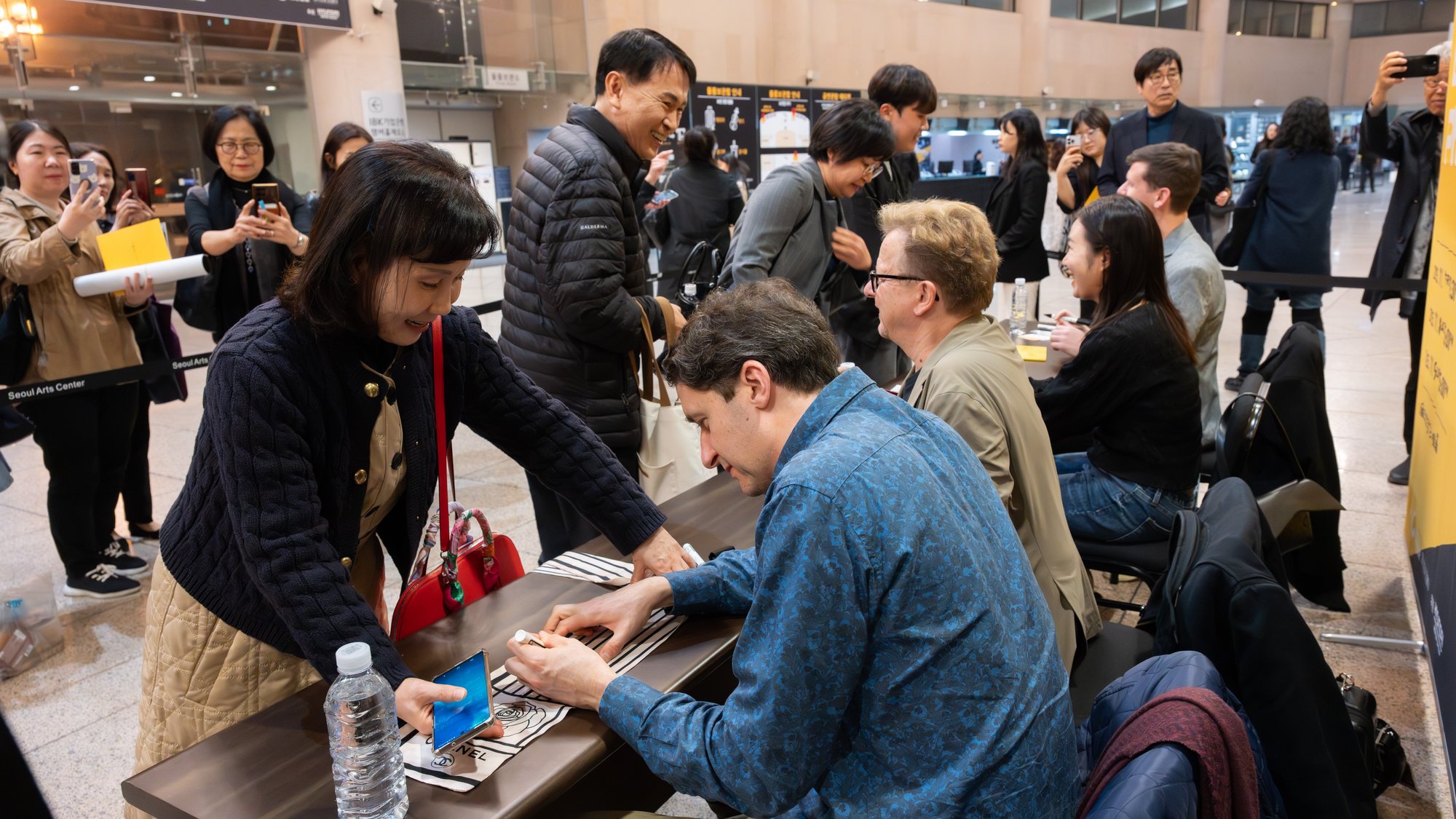 People gather around a table for an autograph session; musicians sign programs for fans, while others wait in line and take photos in a busy indoor venue.