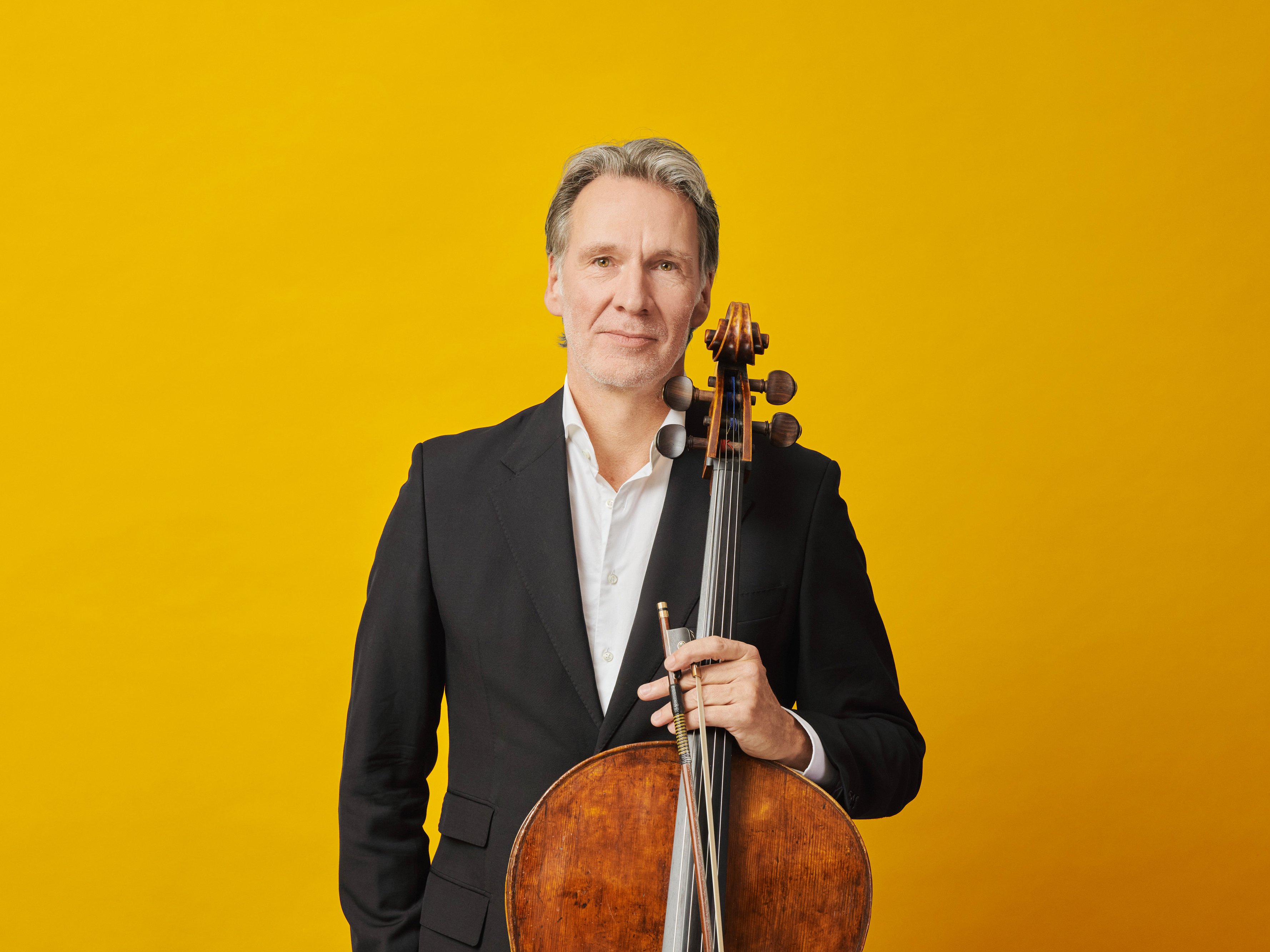 Ein Mann in einem schwarzen Anzug steht mit einem Cello in der Hand vor einem leuchtend gelben Hintergrund und schaut mit neutralem Blick direkt in die Kamera.