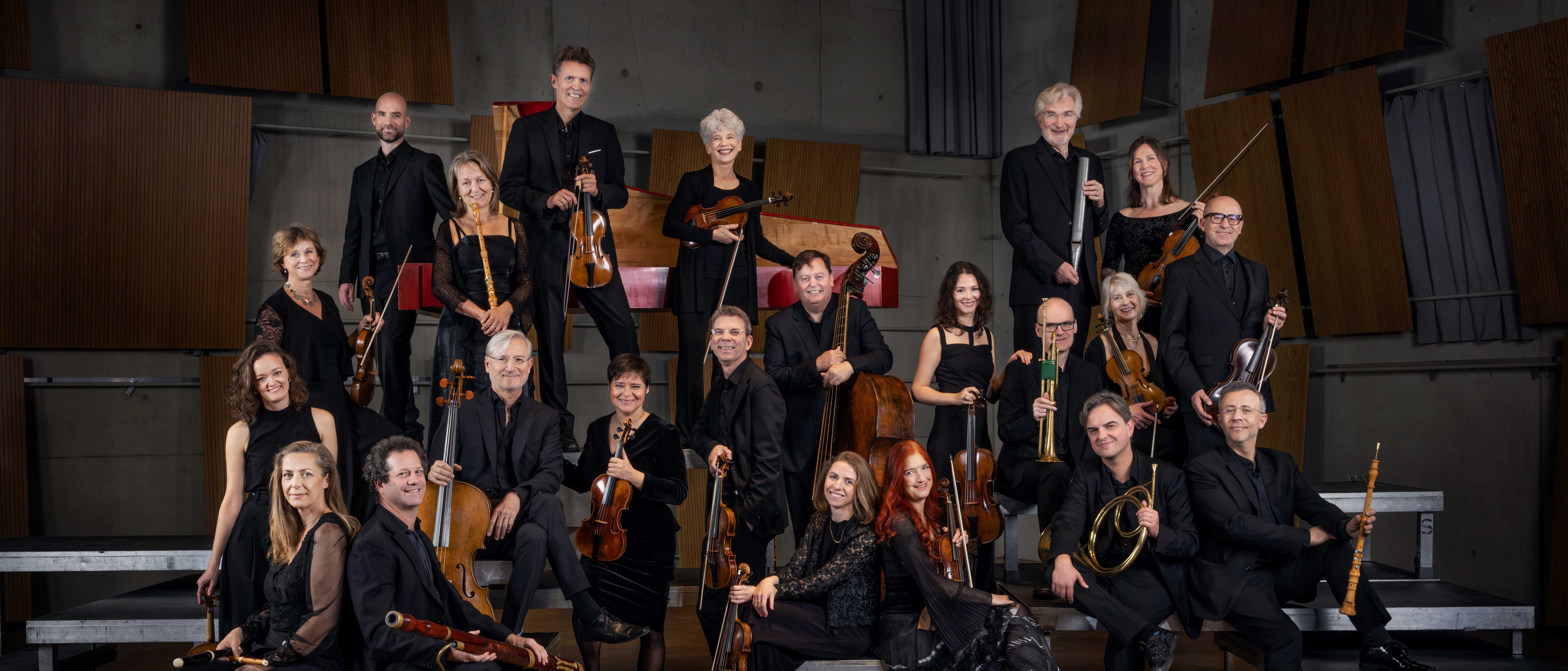 Eine Gruppe von 21 schwarz gekleideten Musikern mit verschiedenen Streich- und Blasinstrumenten posiert gemeinsam auf den Rängen einer modernen Konzerthalle.