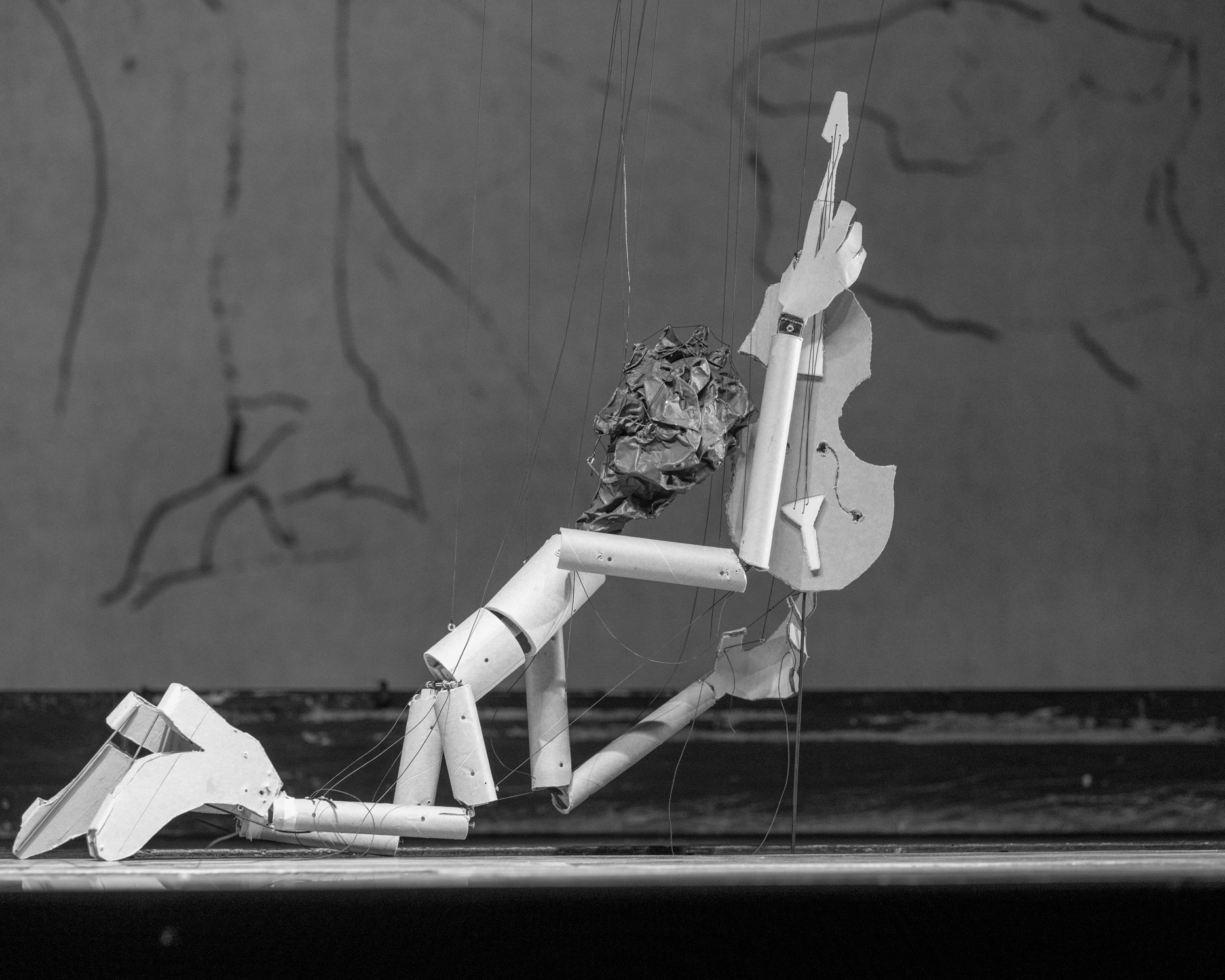 Eine Marionette hält, auf den Knien abgestützt, ein Streichinstrument in die Höhe.