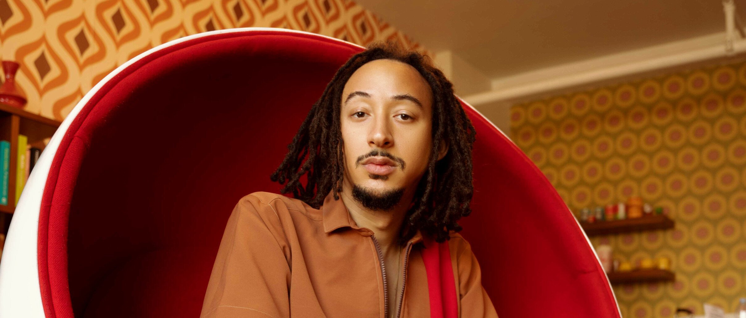 A person with dreadlocks sits confidently in a retro, red and white egg chair, wearing a brown outfit and black boots, in a room with bold geometric wallpaper and a mid-century modern vibe.