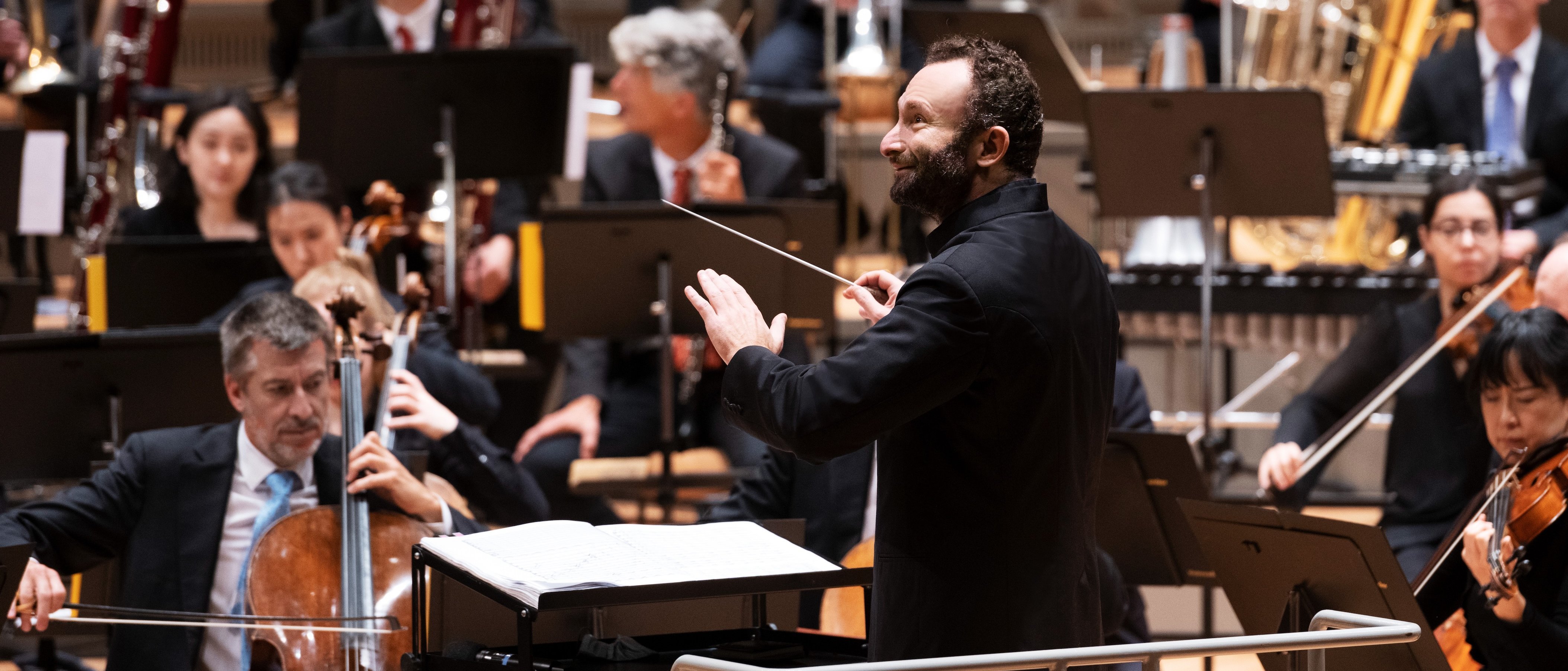 Ein Dirigent leitet ein Orchester während einer Aufführung; die Musiker spielen verschiedene Instrumente, darunter Streicher und Blechbläser, während sie die Noten auf Ständern lesen.