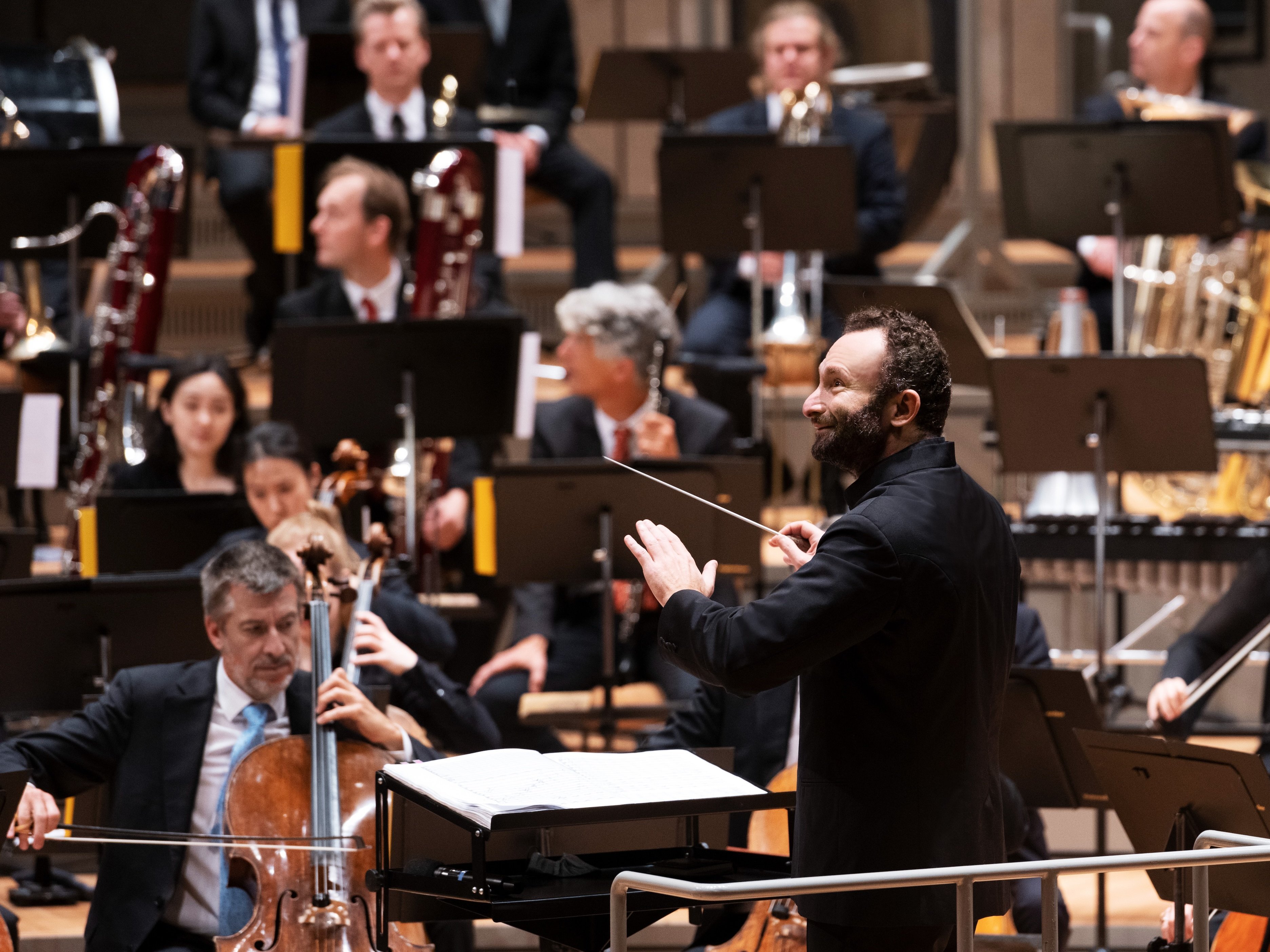 Ein Dirigent leitet ein Orchester während einer Aufführung; die Musiker spielen verschiedene Instrumente, darunter Streicher und Blechbläser, während sie die Noten auf Ständern lesen.