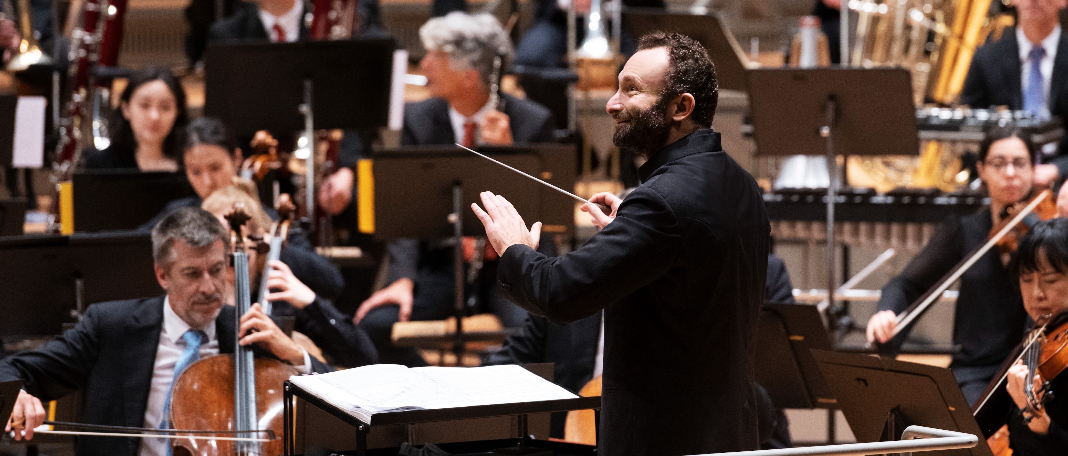 Ein Dirigent leitet ein Orchester während einer Aufführung; die Musiker spielen verschiedene Instrumente, darunter Streicher und Blechbläser, während sie die Noten auf Ständern lesen.