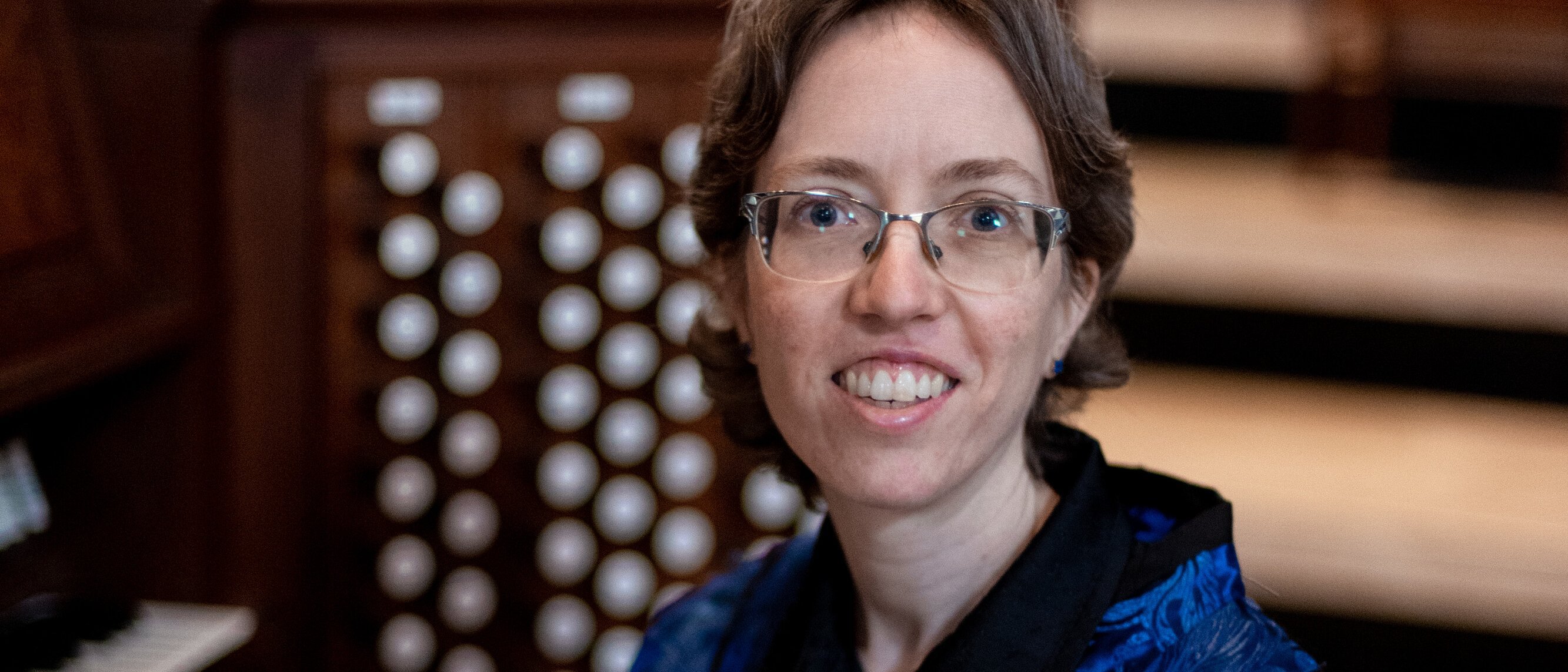 In einem holzgetäfelten Raum sitzt Isabelle Demers mit kurzen braunen Haaren und Brille an einem Orgel-Keyboard, lächelt und trägt eine blau gemusterte Jacke.