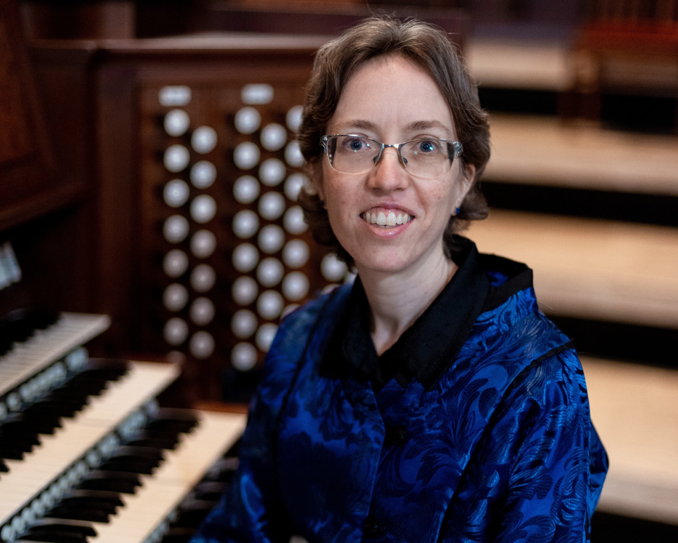 In einem holzgetäfelten Raum sitzt Isabelle Demers mit kurzen braunen Haaren und Brille an einem Orgel-Keyboard, lächelt und trägt eine blau gemusterte Jacke.