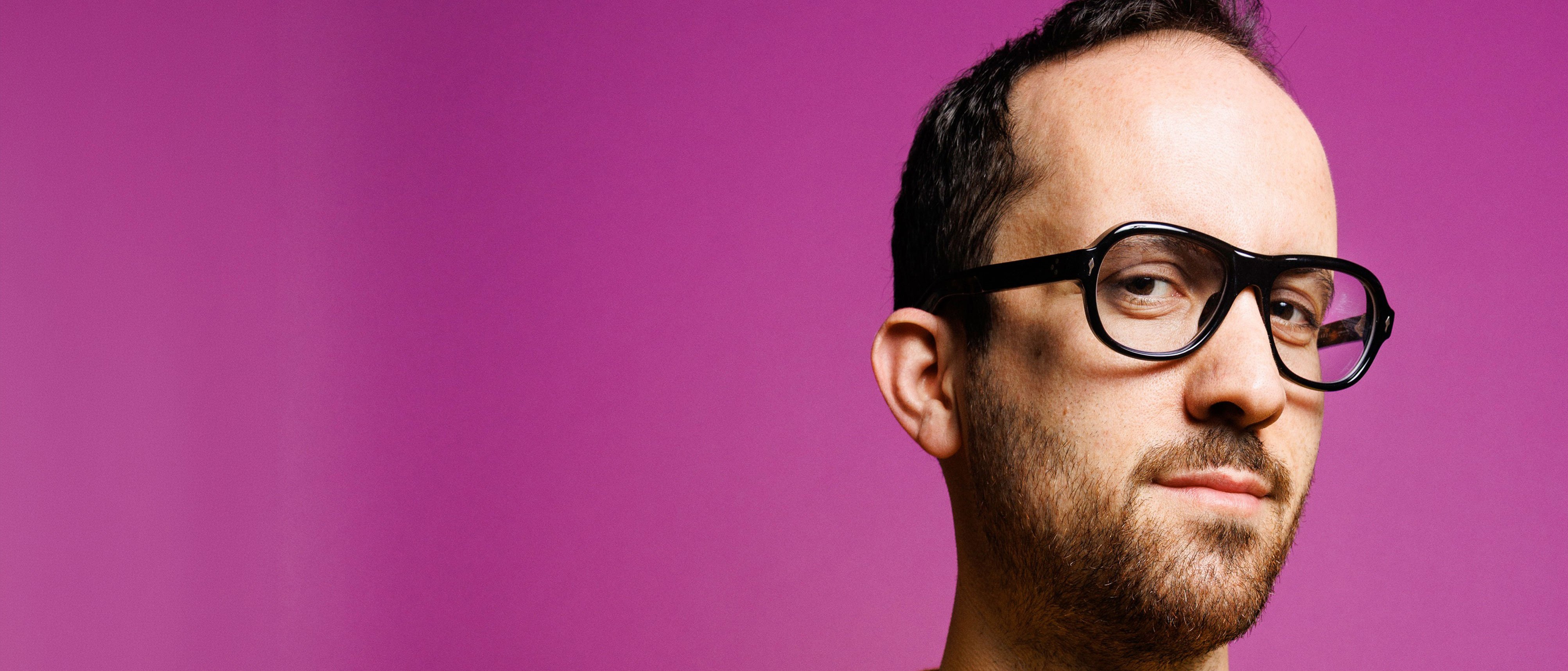 Ein Mann mit kurzem Bart, der eine große schwarze Brille und einen gelben Pullover trägt, blickt mit einem leichten Lächeln in die Kamera, vor einem einfarbigen rosa Hintergrund.
