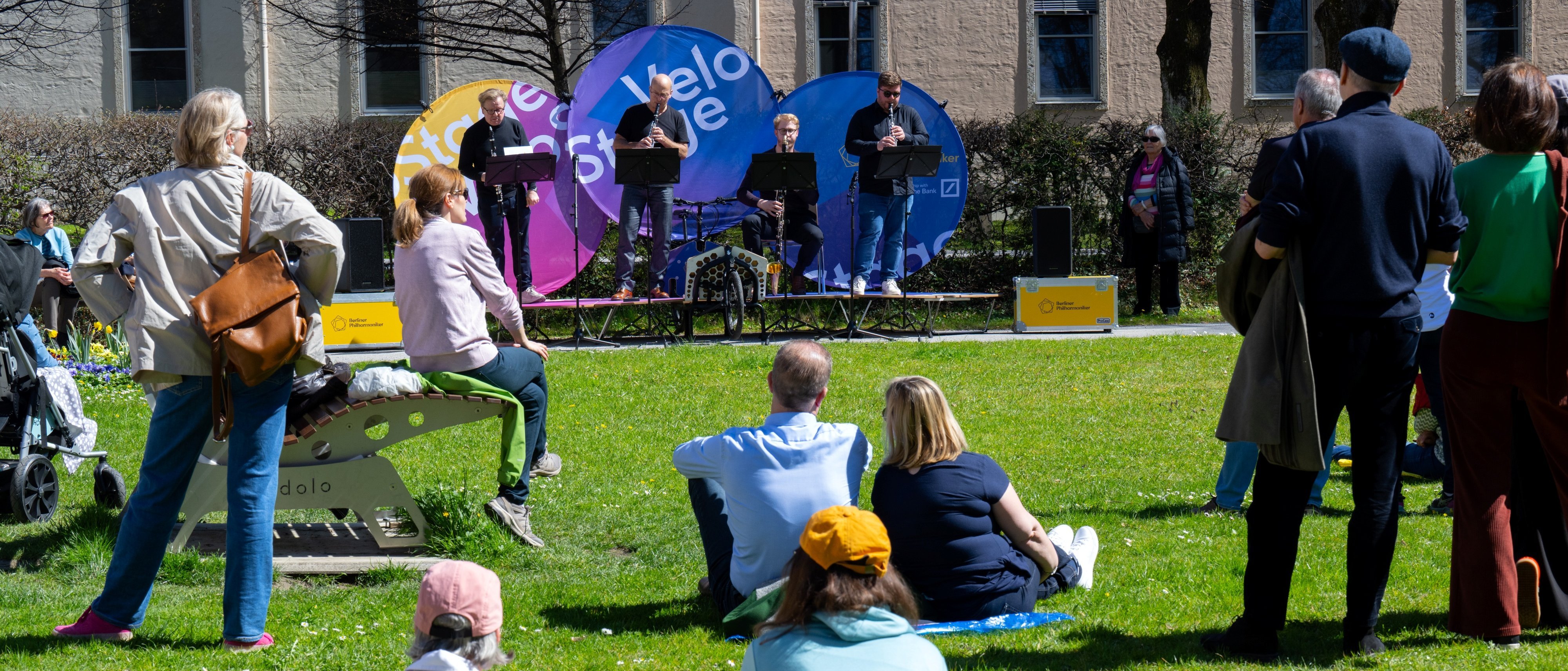 Menschen sitzen und stehen auf einer Wiese und beobachten eine Fahrradbühne, auf der vier Musiker auftreten. Bunte Fahnen und Bäume umgeben das Gelände, und ein paar Leute genießen den sonnigen Tag.