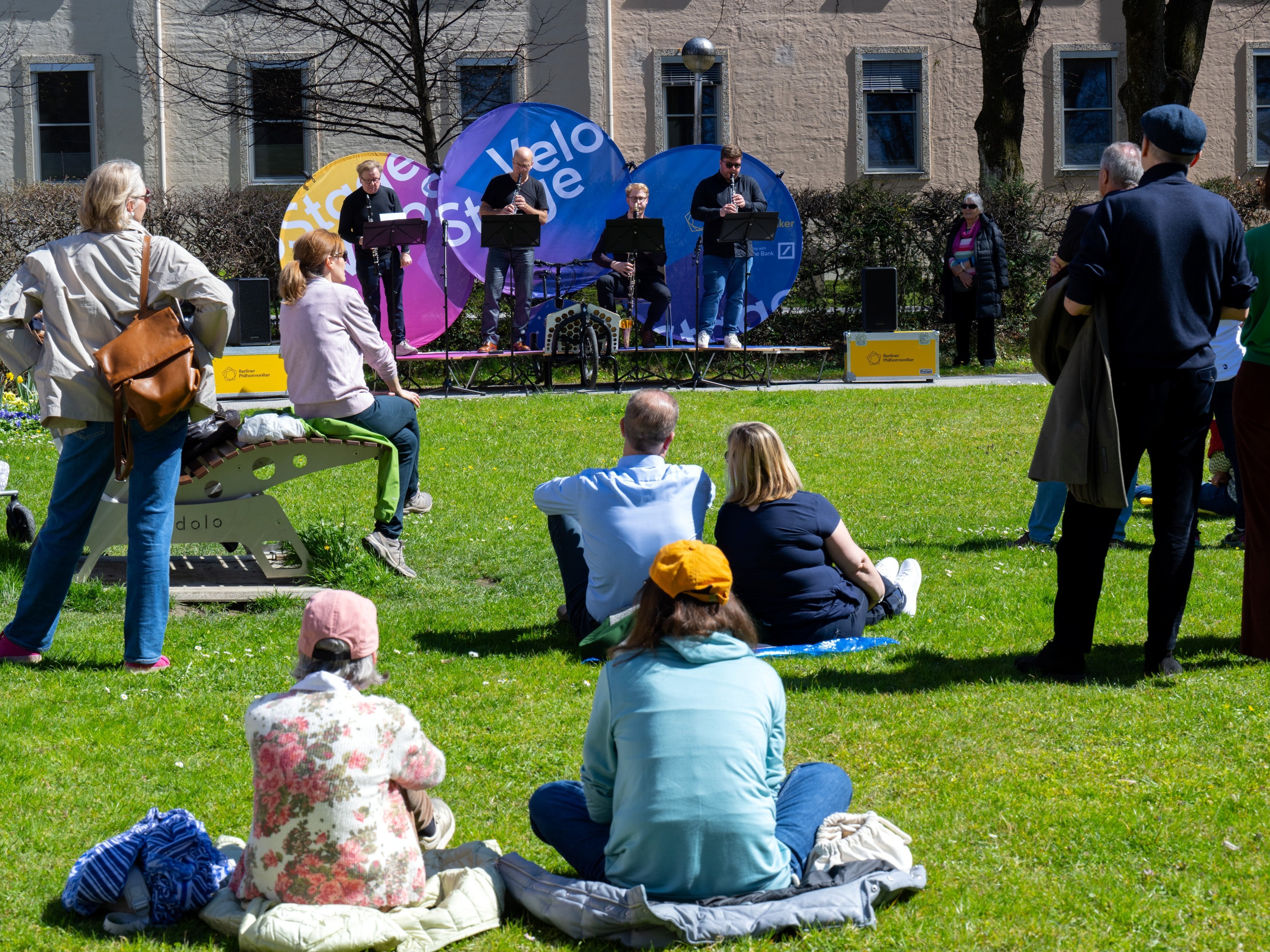 Menschen sitzen und stehen auf einer Wiese und beobachten eine Fahrradbühne, auf der vier Musiker auftreten. Bunte Fahnen und Bäume umgeben das Gelände, und ein paar Leute genießen den sonnigen Tag.