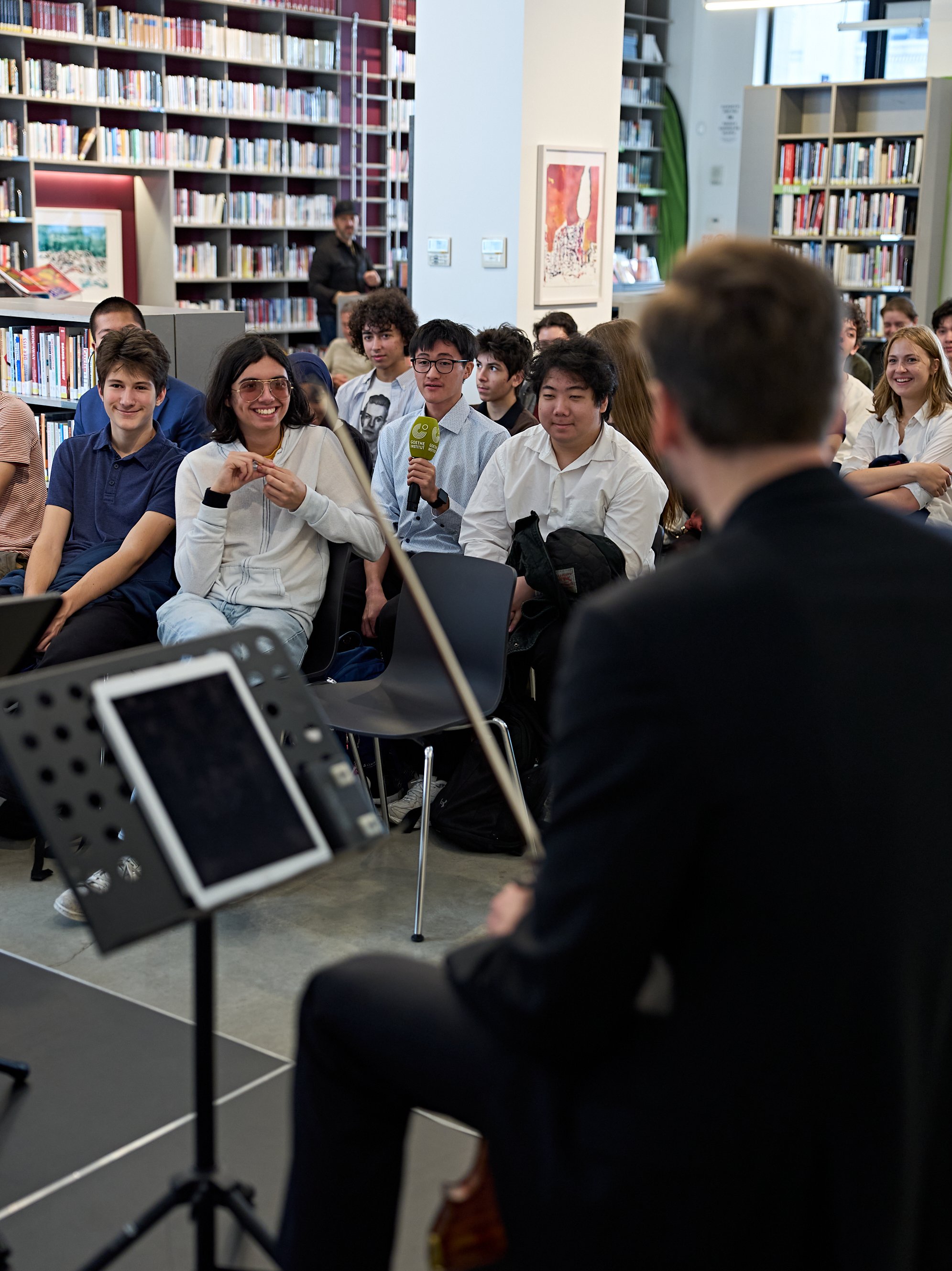 Ein Musiker spielt Saiteninstrumente vor einem Publikum aus Schülern, die in einer Bibliothek sitzen.