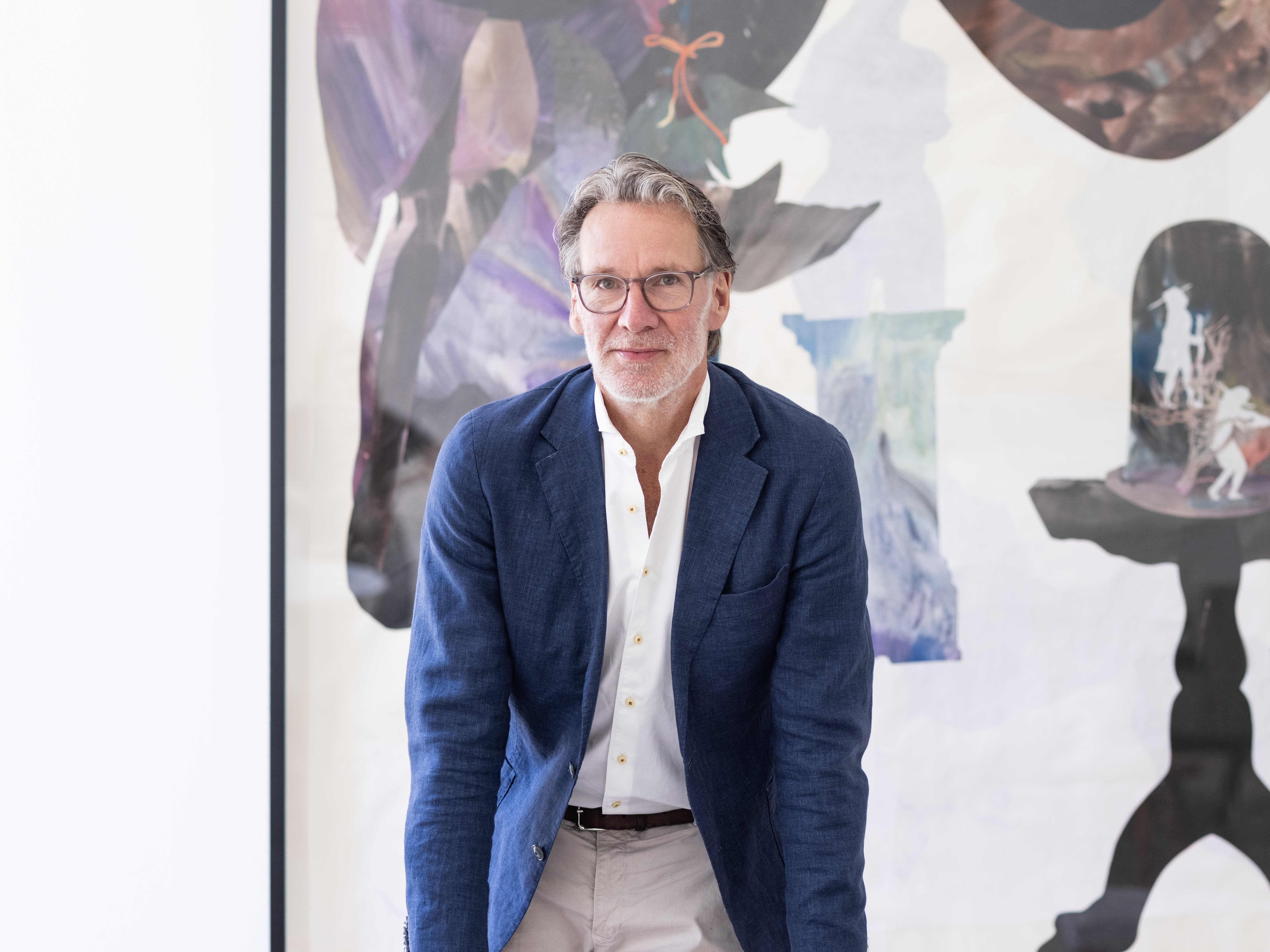 Ein Mann in einem blauen Blazer lehnt sich an einen hellen Stuhl und steht vor einem großen abstrakten Gemälde mit dunklen und hellen Formen in einem hellen, modernen Raum.