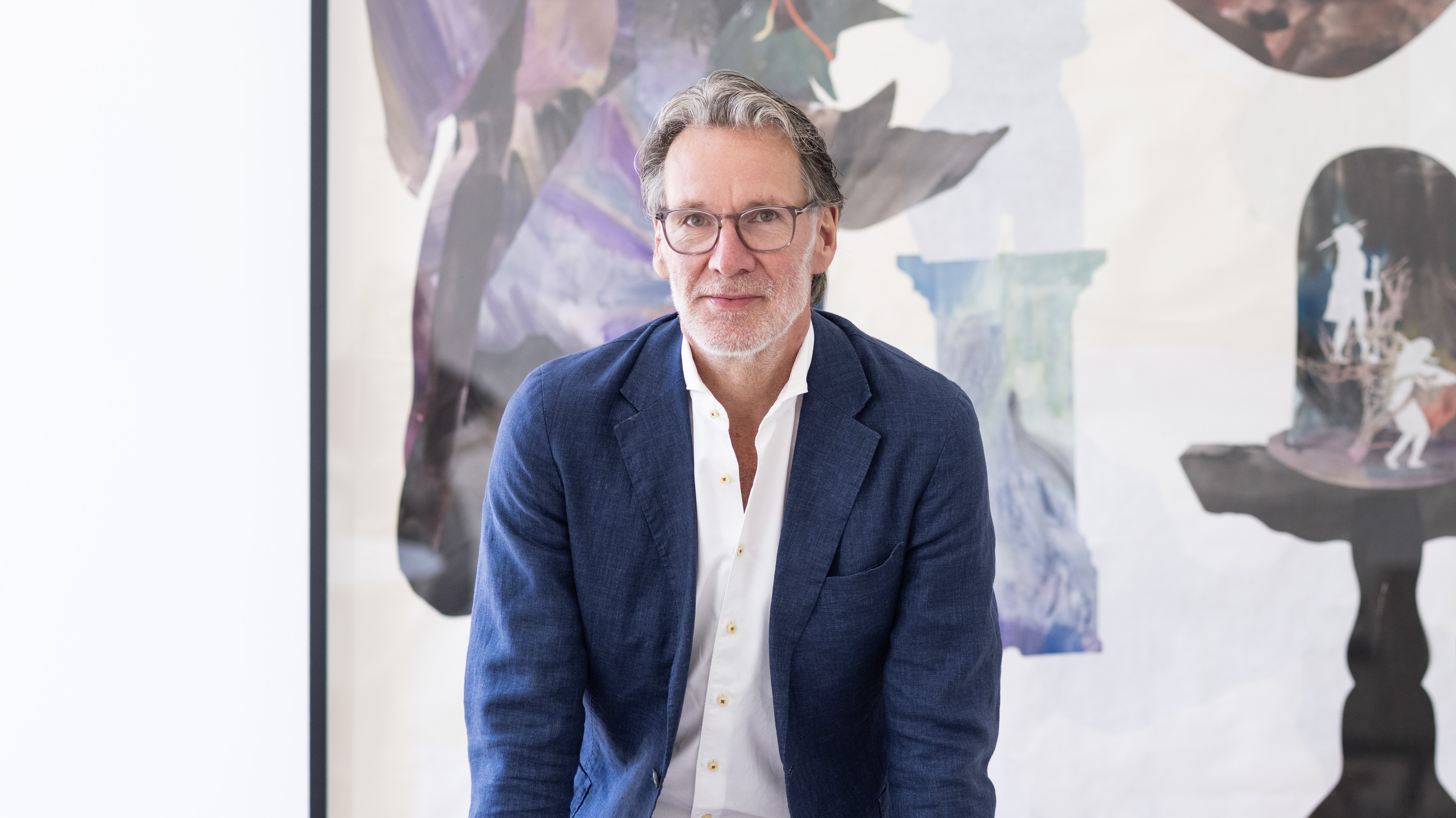 Ein Mann in einem blauen Blazer lehnt sich an einen hellen Stuhl und steht vor einem großen abstrakten Gemälde mit dunklen und hellen Formen in einem hellen, modernen Raum.