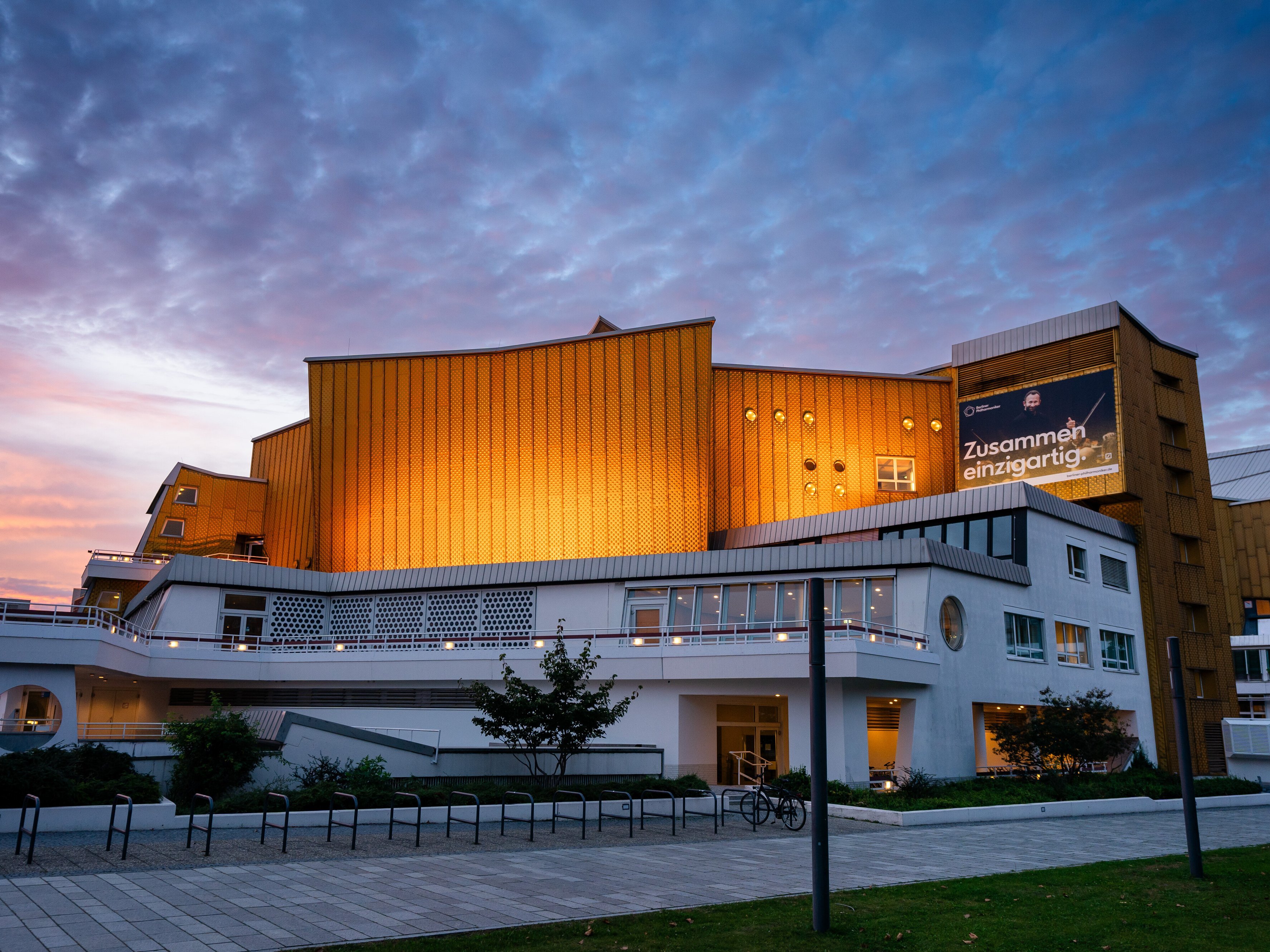 Die Philharmonie Berlin mit goldenen Paneelen und weißen unteren Stockwerken wird in der Abenddämmerung beleuchtet, mit einem bewölkten Himmel im Hintergrund. Auf einem großen Banner steht auf Deutsch »Zusammen einzigartig!.